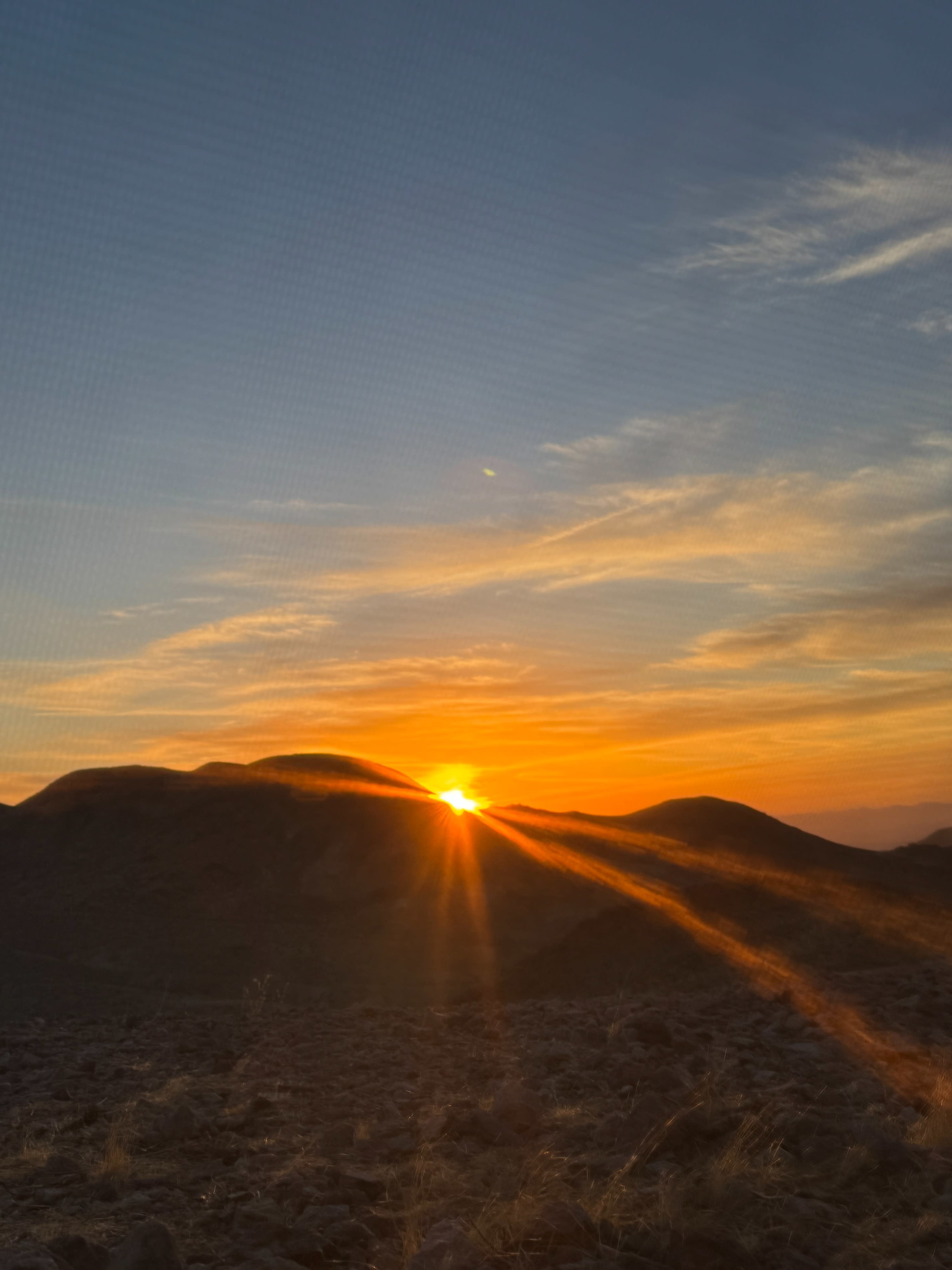The sun rises over a mountainous landscape, casting rays of light across the rocky terrain.