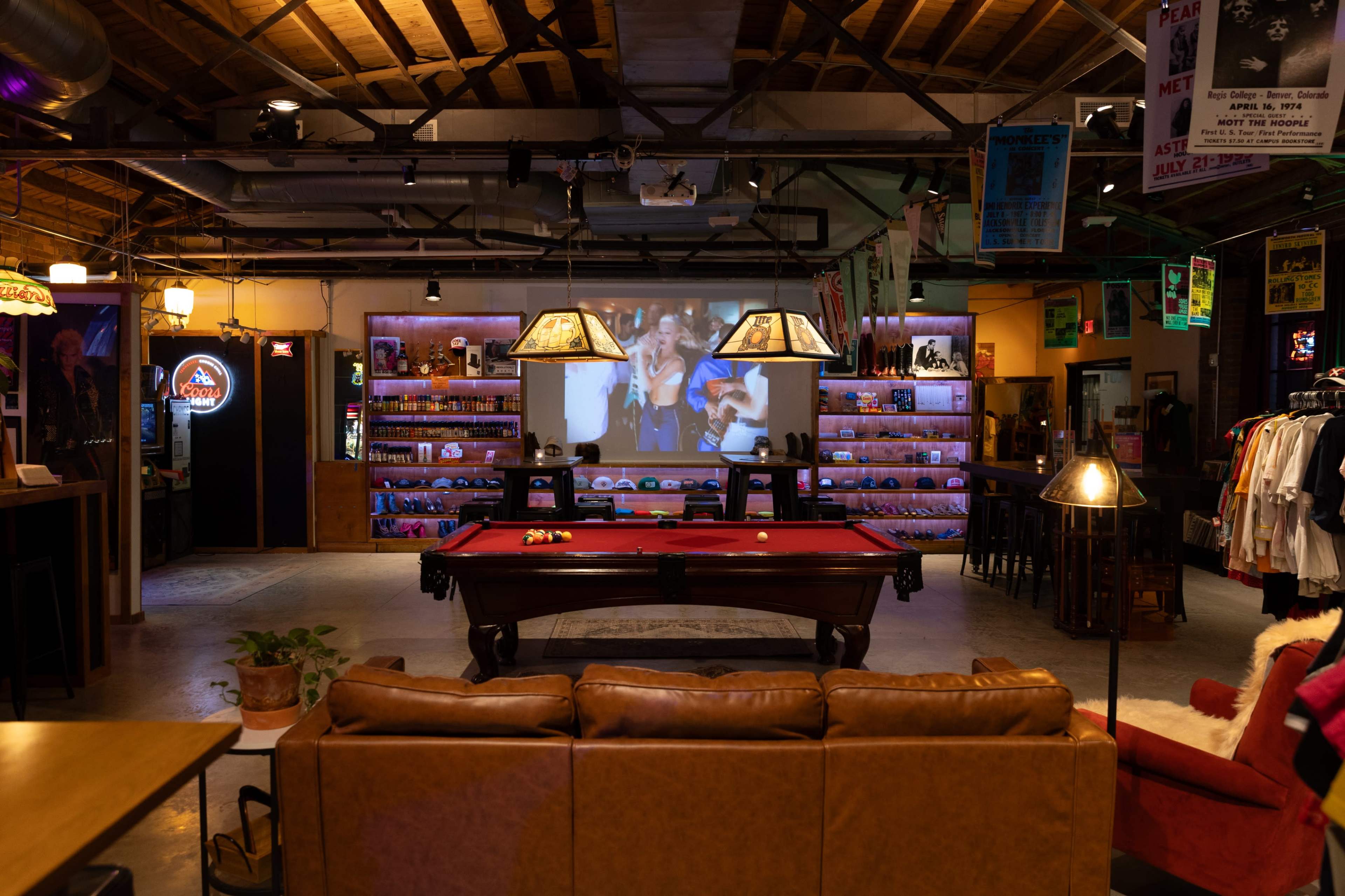 The scene features a spacious lounge with a billiards table in the foreground, surrounded by various seating areas and shelves displaying items, while a large screen shows a video at the back.