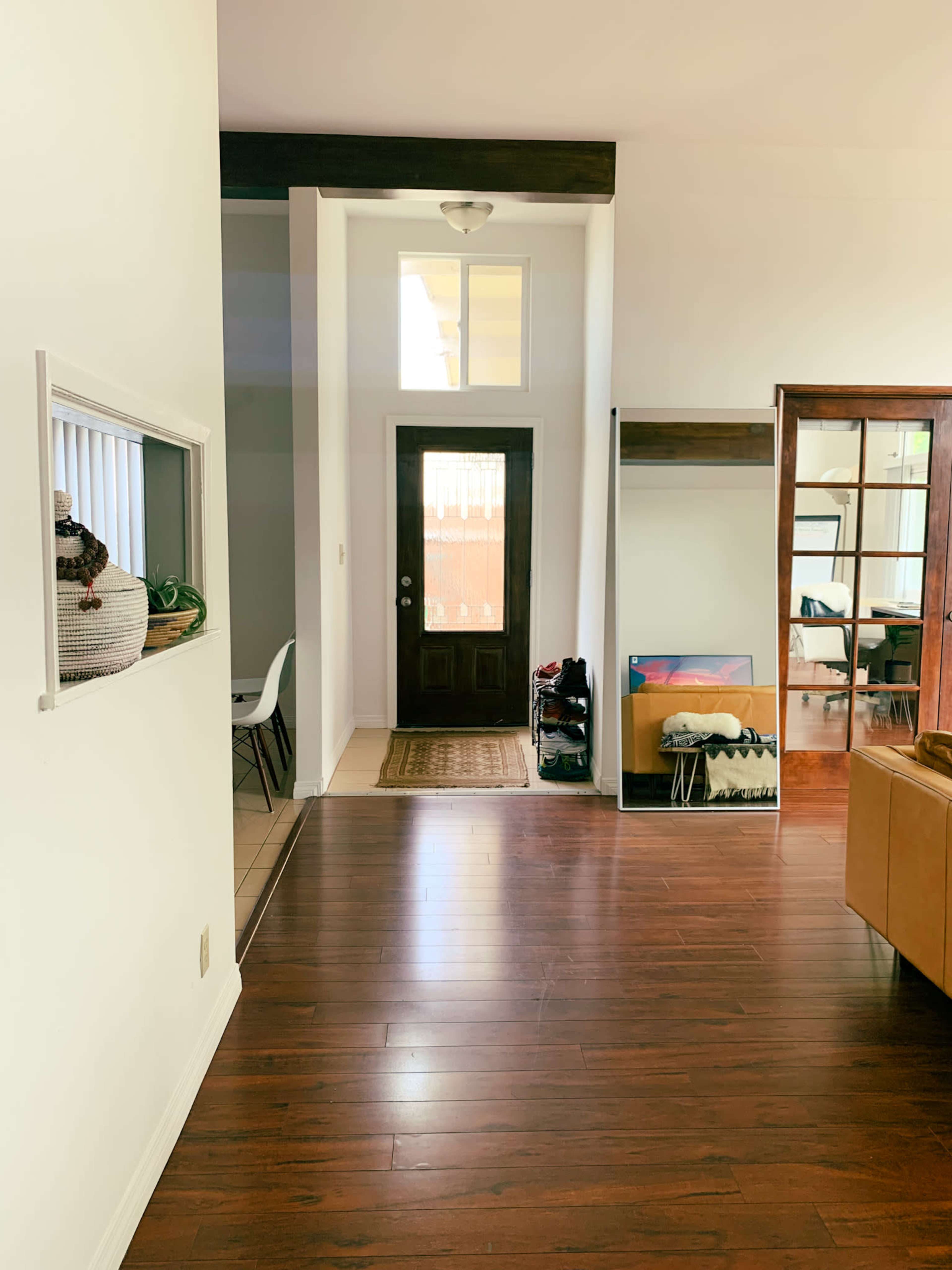 A hallway with wooden flooring, leading to a front door, with a mirror and some decorative elements along the walls.