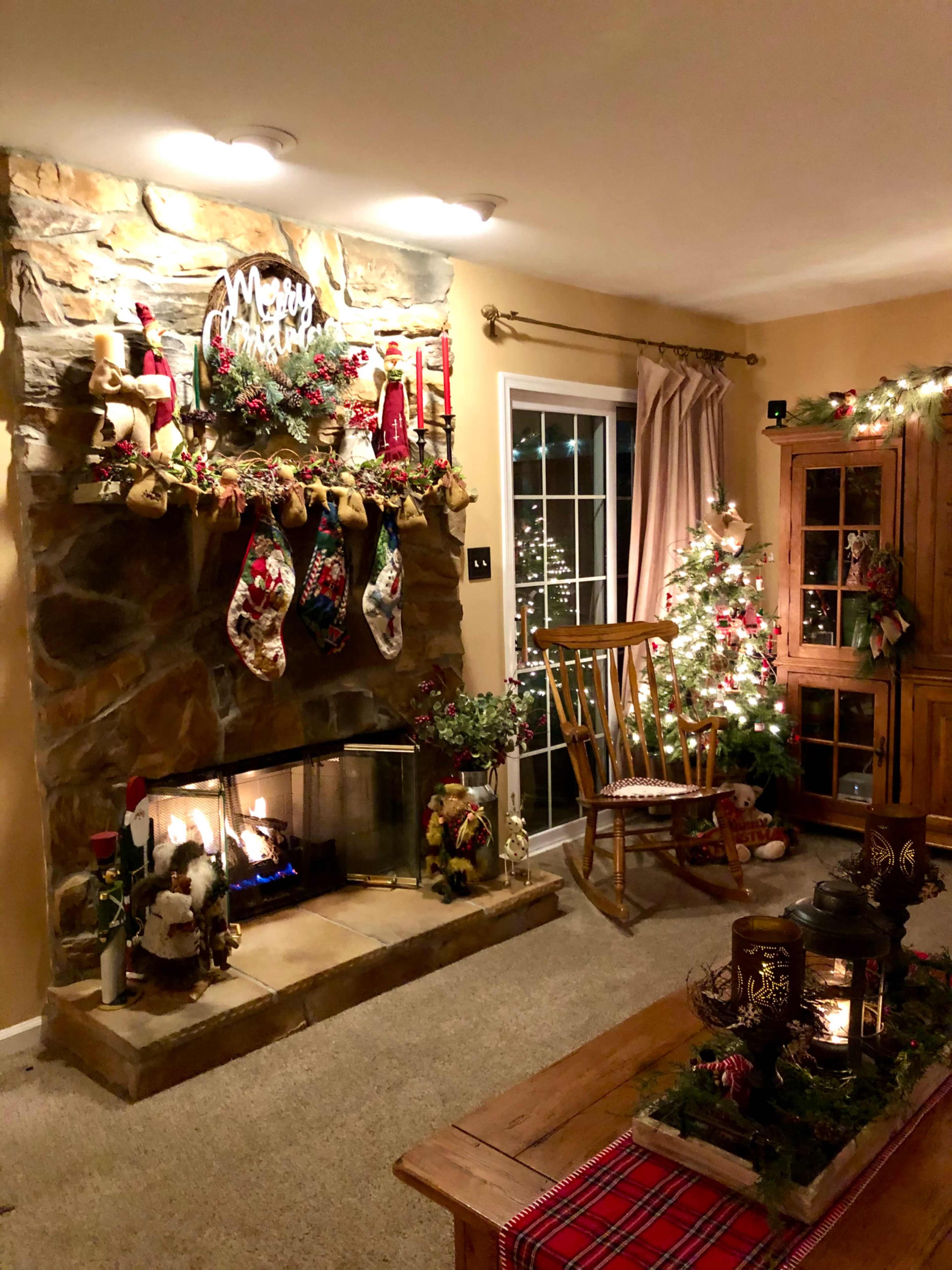 The image shows a cozy living room decorated for Christmas, featuring a stone fireplace with stockings, a small Christmas tree, and festive lights.