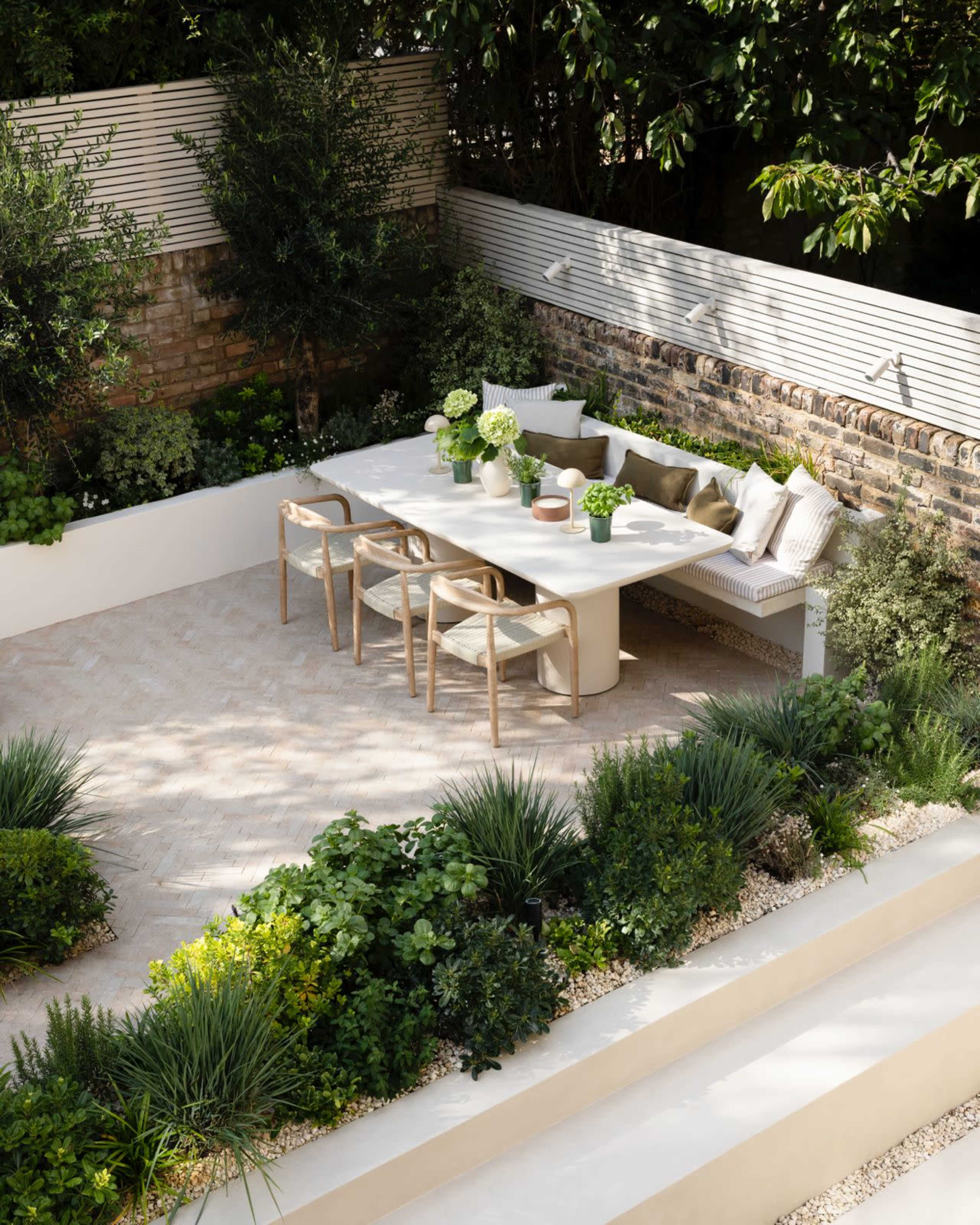 A modern outdoor dining area features a long white table surrounded by wooden chairs, with potted plants and greenery arranged around the space.
