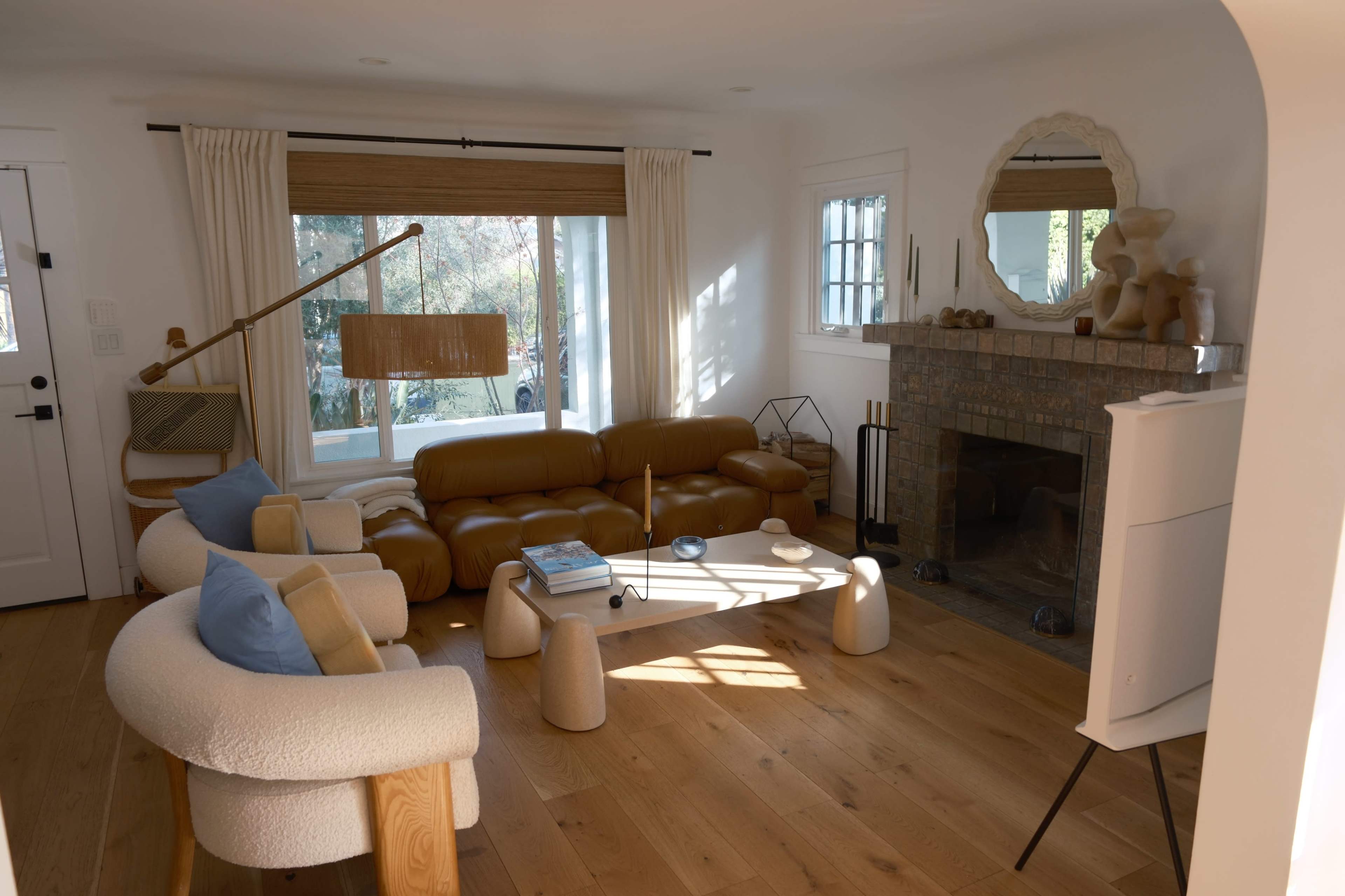 The image shows a bright and modern living room featuring a leather sectional sofa, a circular mirror above a fireplace, and light wooden flooring.