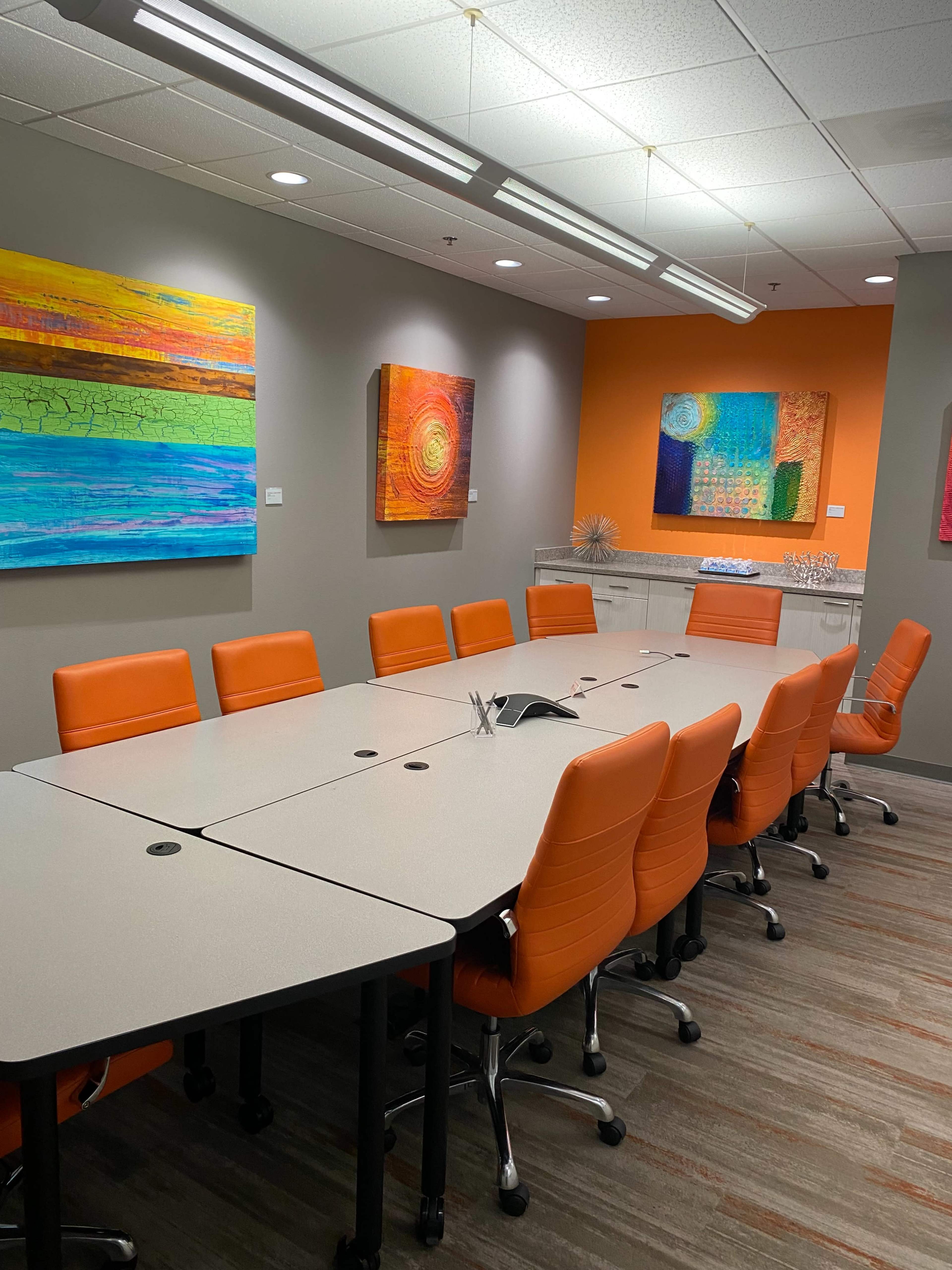 A modern conference room features a long table surrounded by orange chairs, with colorful abstract art displayed on the walls.