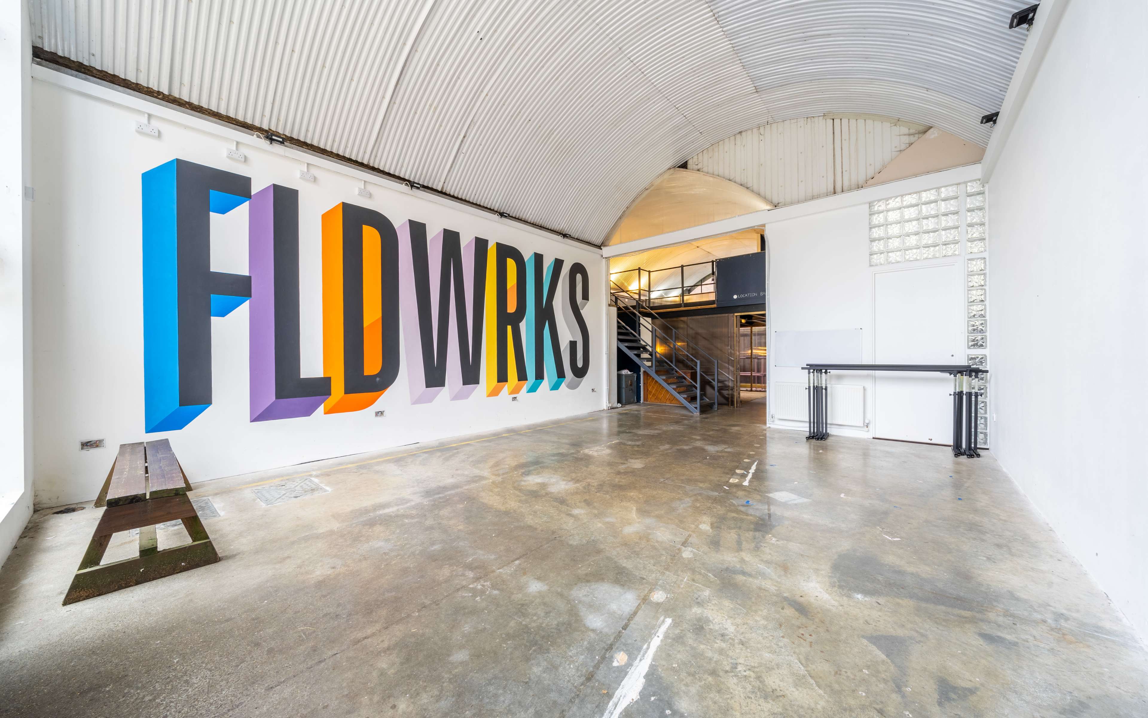 An empty, industrial-style interior with a large mural that reads "FLDWRKS" on one wall and a staircase leading to an upper level.