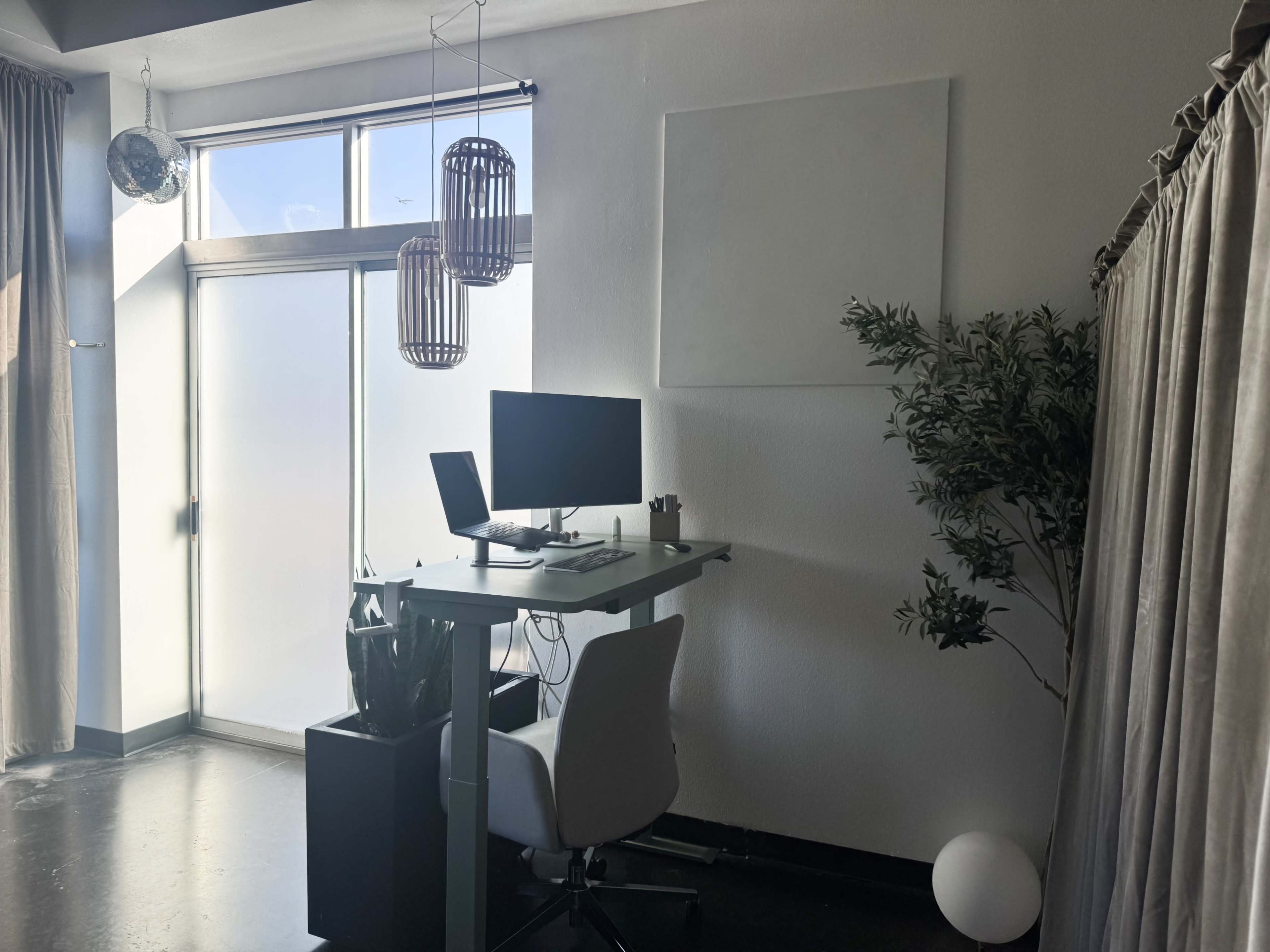 The image shows a minimalistic home office setup featuring a desk with a computer, a plant, and large windows allowing light to enter the space.