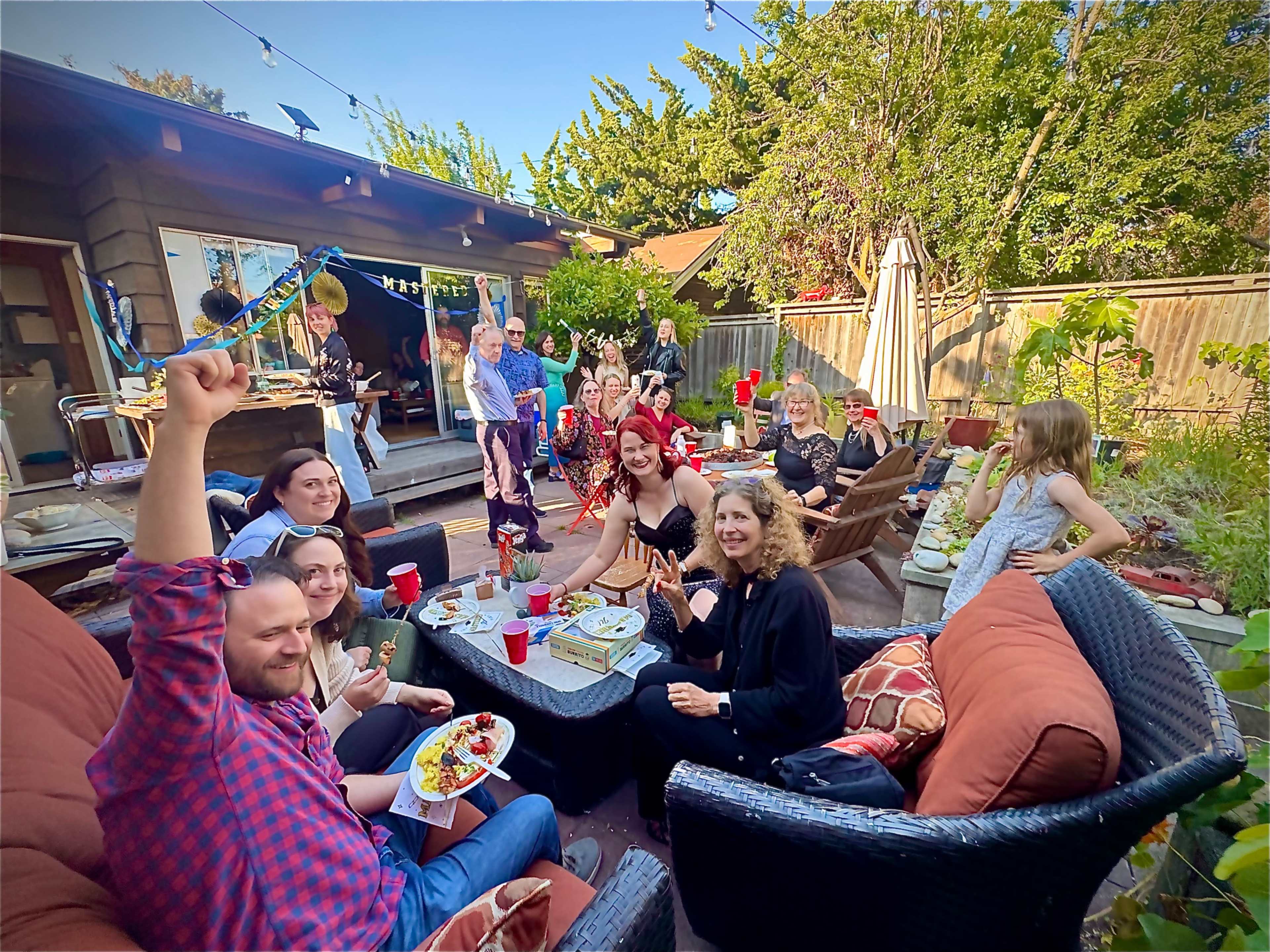 A group of people gathers in a backyard for a social event, sitting around a patio area with food and drinks, while others mingle in the background.