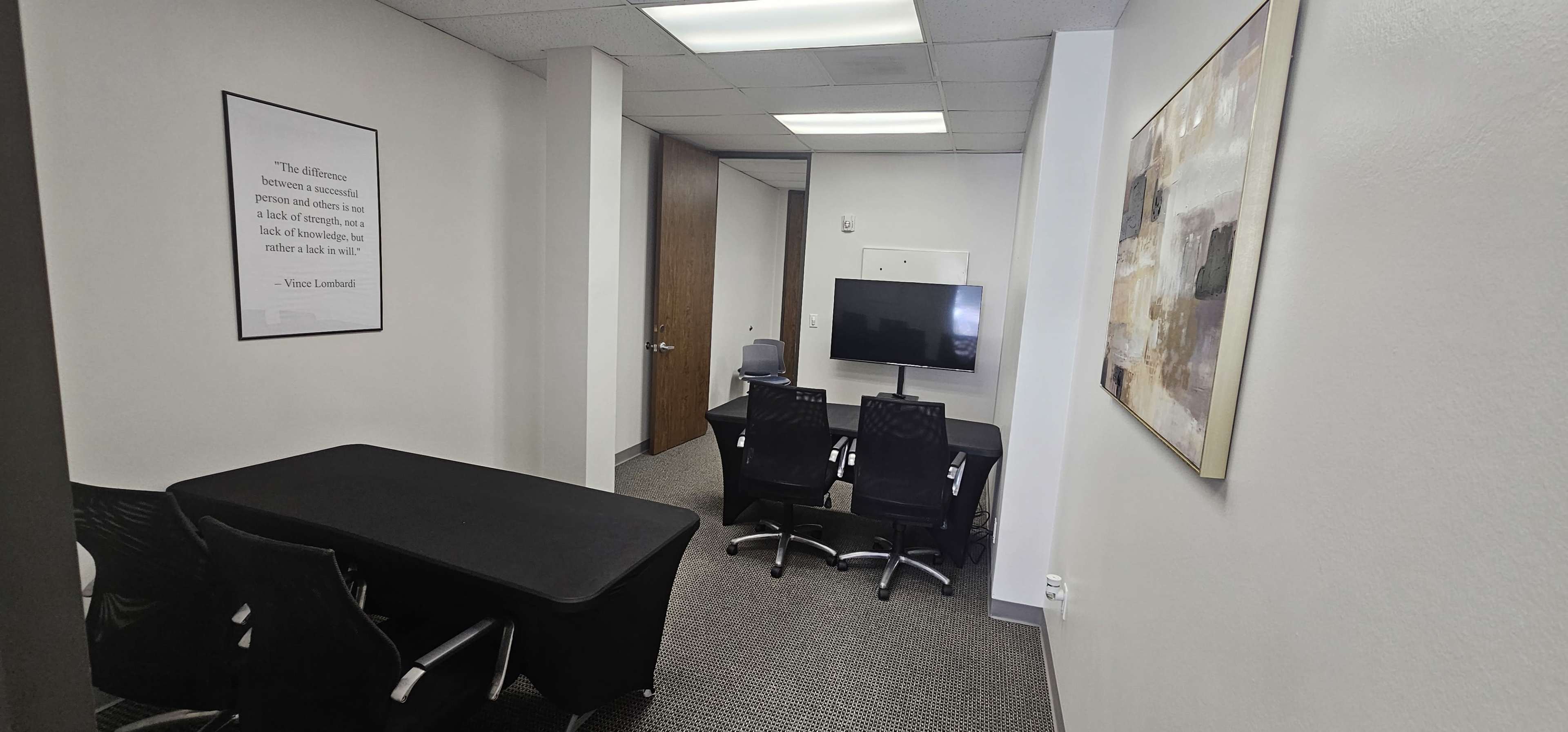 The image shows a small office space with a table, chairs, and a television mounted on the wall, along with a framed artwork.