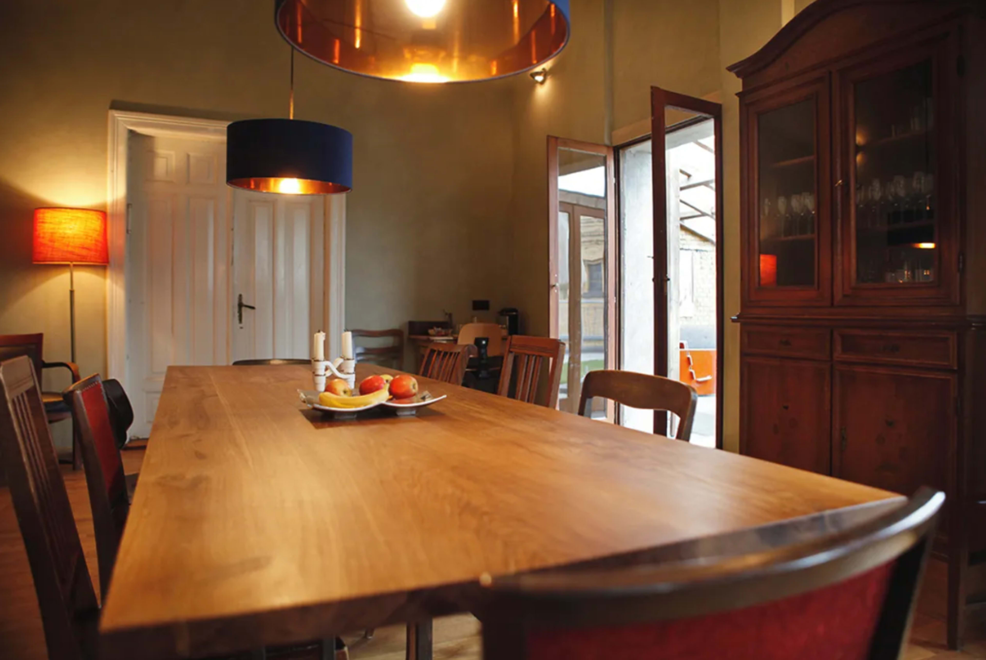 A large wooden dining table is set in a well-lit room with a cabinet, colorful lamps, and open doors leading to an outdoor area.