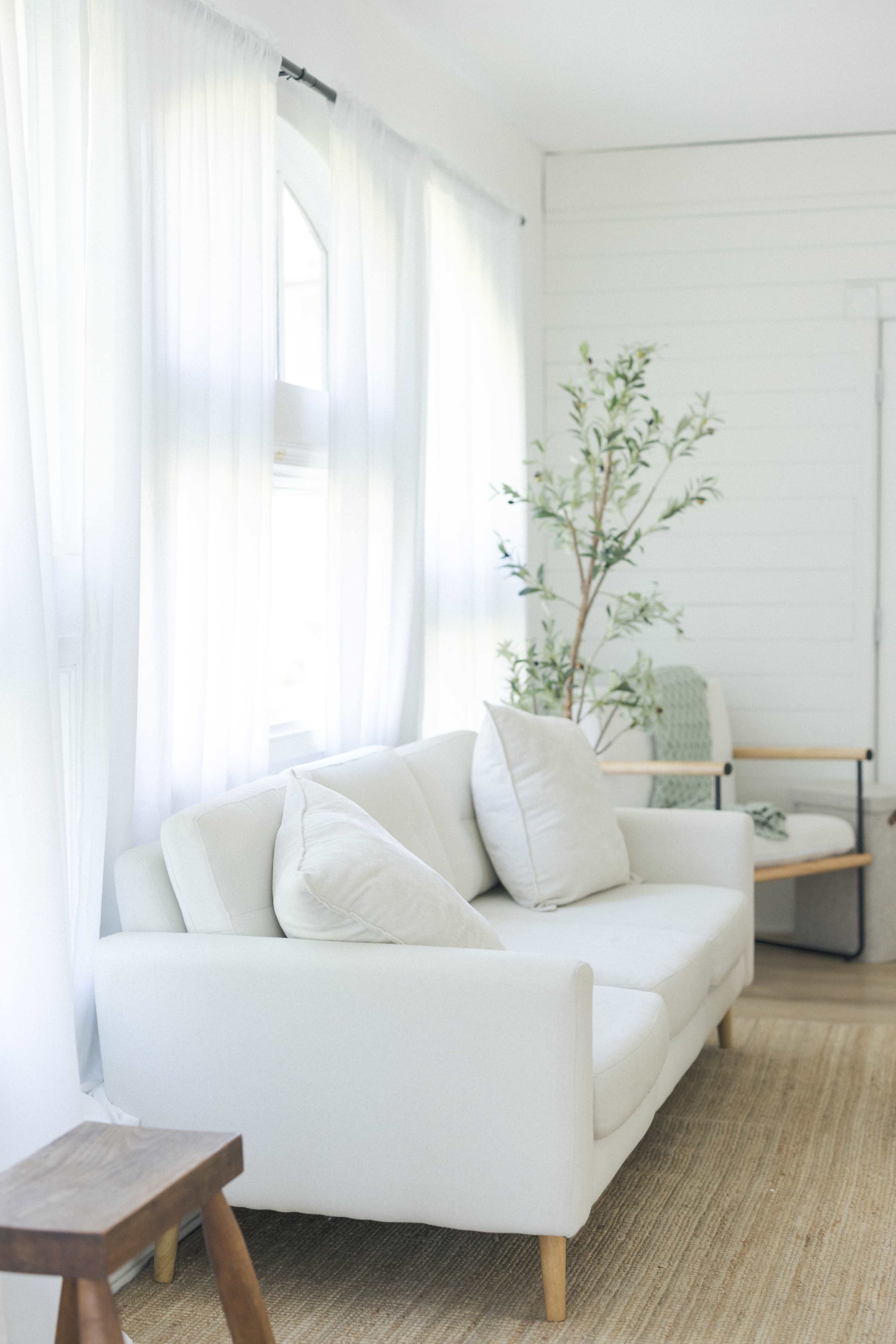 A bright living room features a white sofa with two pillows, a wooden side table, and a potted plant beside a chair.