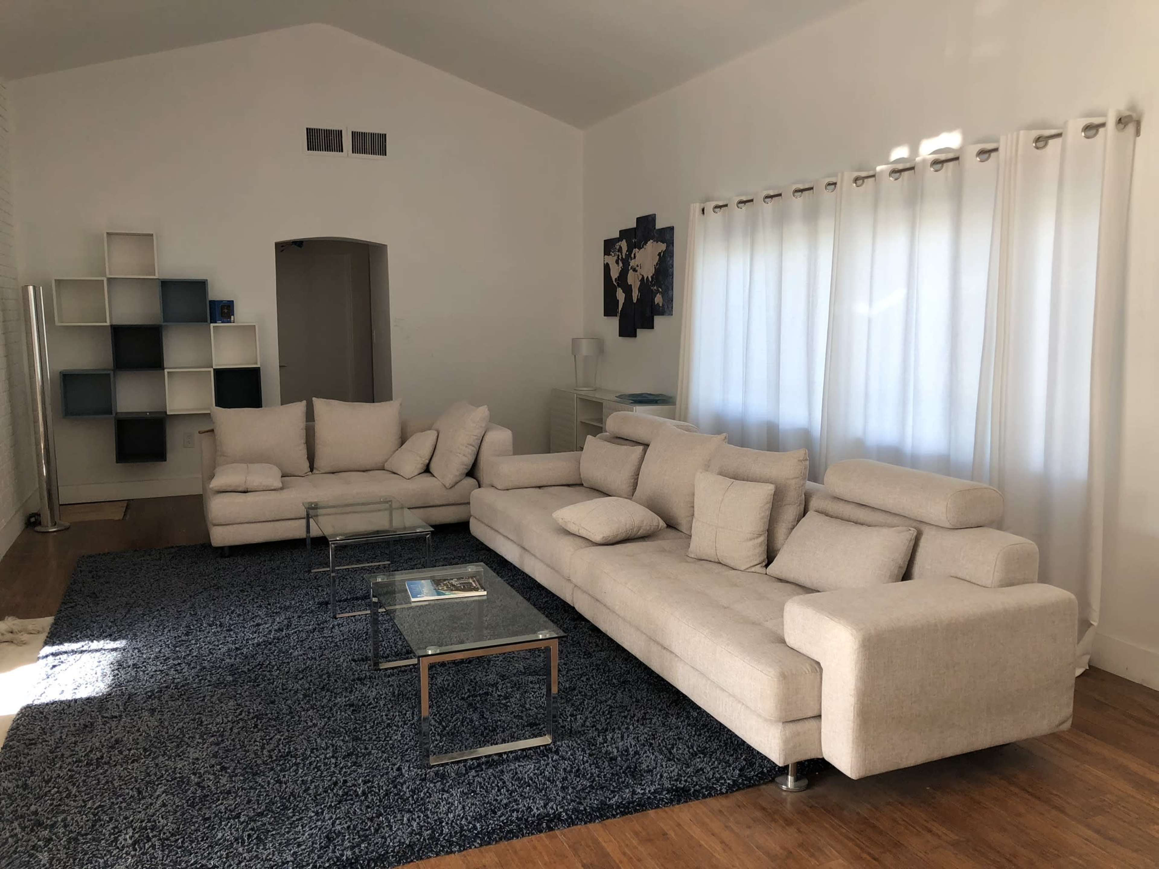 A modern living room featuring a large light-colored sectional sofa, a glass coffee table, and a textured dark rug, with natural light streaming through sheer curtains.