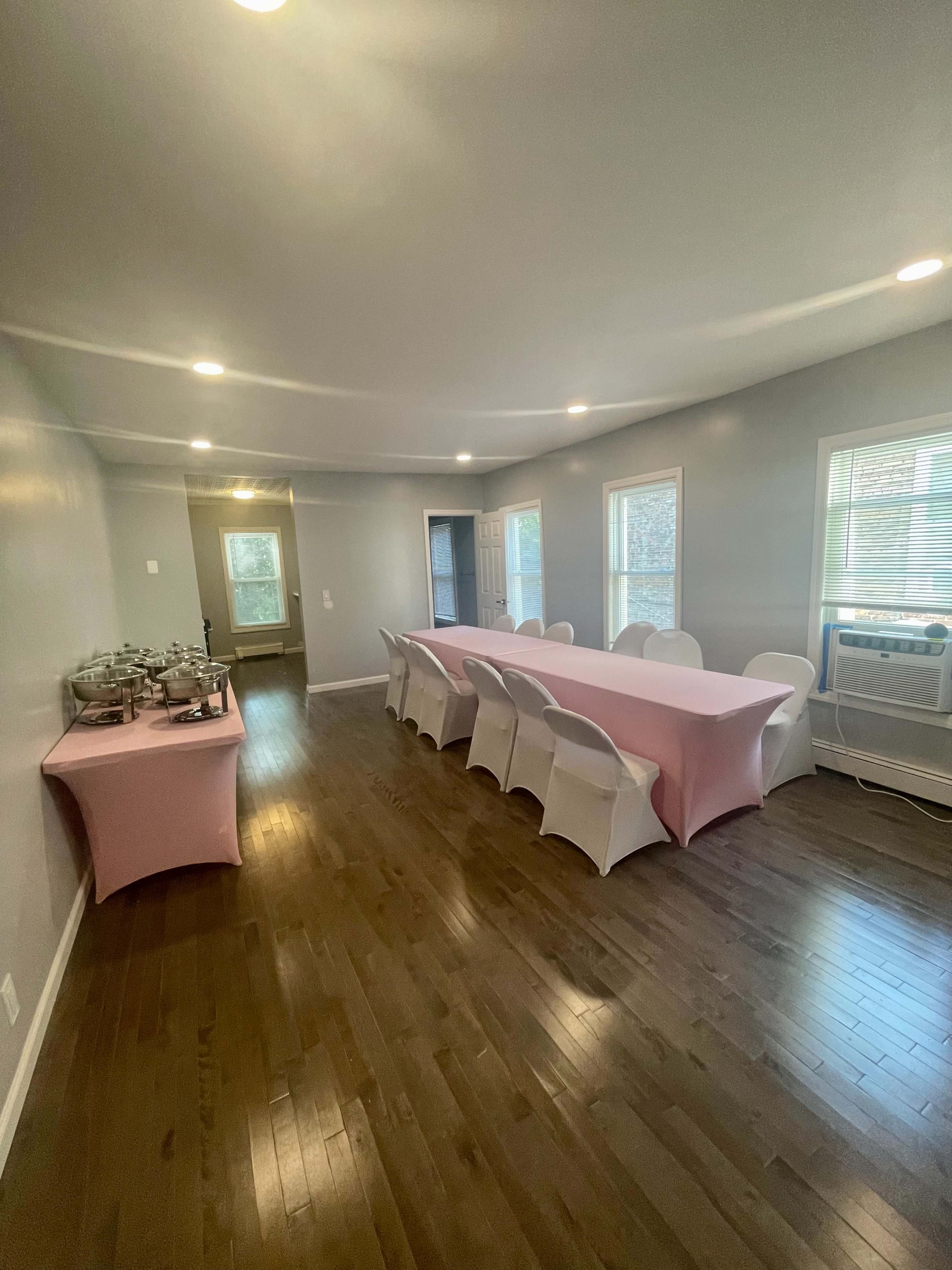 The room features a long table covered with a pink tablecloth surrounded by white chairs, with large windows allowing natural light to enter.
