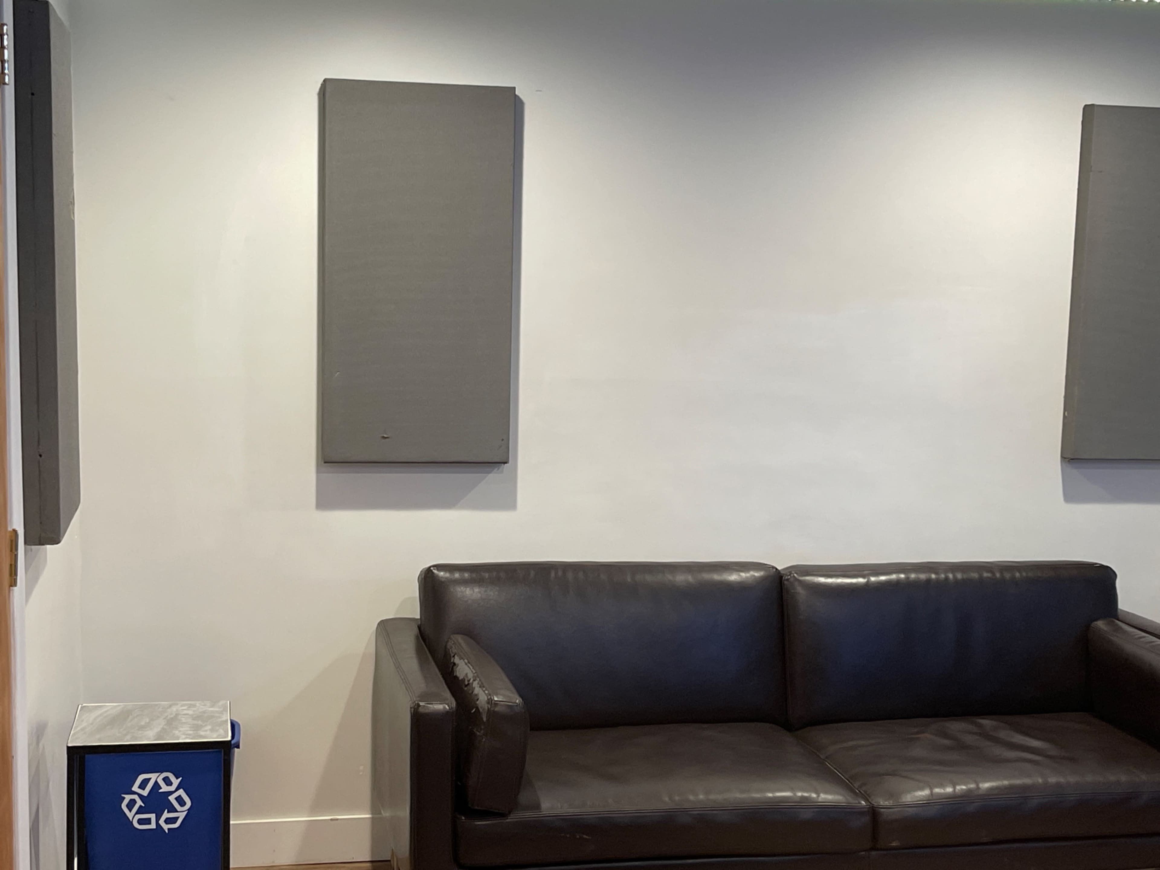 A black leather couch is positioned against a plain wall, with two soundproofing panels mounted above it and a recycling bin nearby.