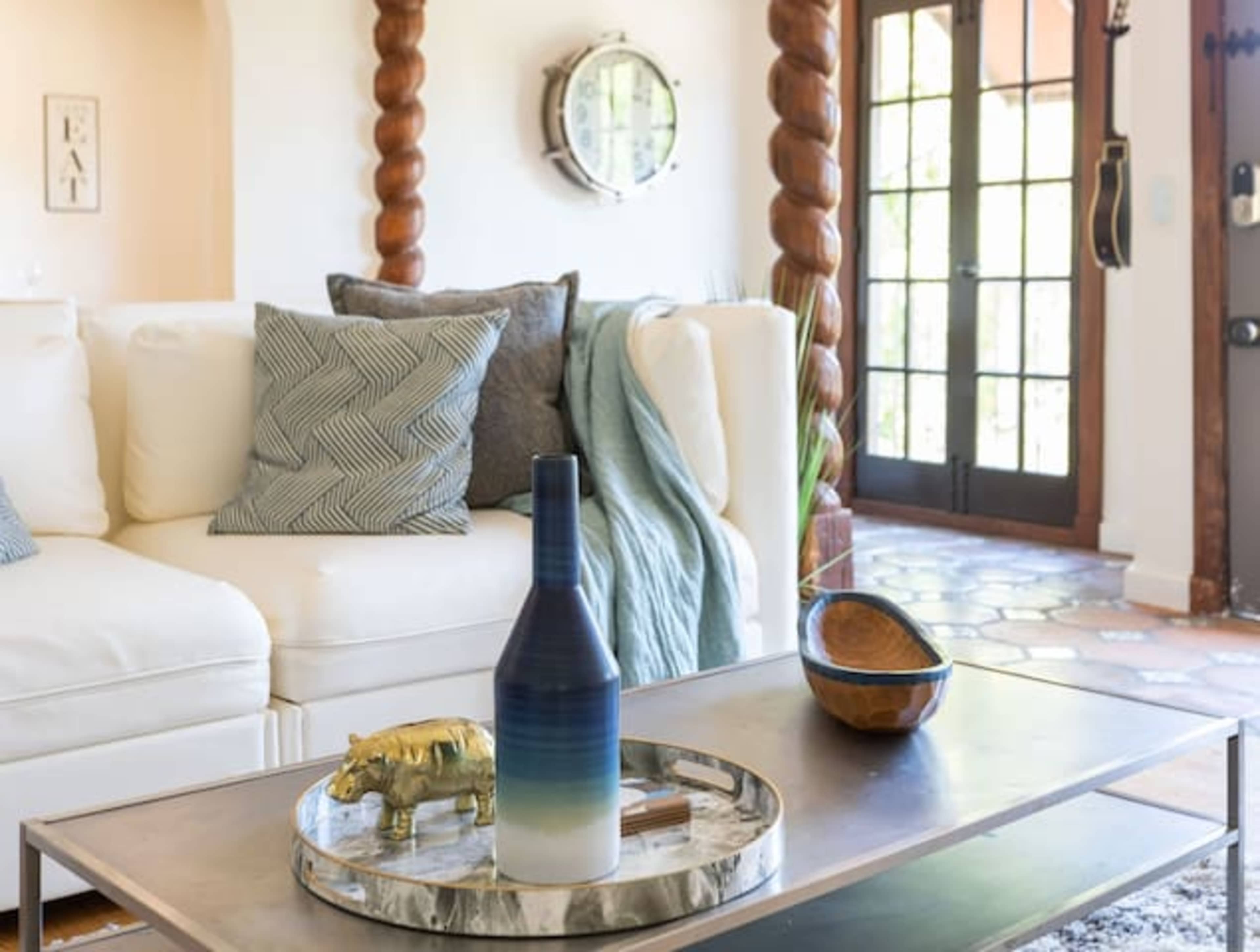 A modern living room features a white sofa adorned with patterned pillows, a glass coffee table with a decorative tray, and a doorway leading to an outdoor area.