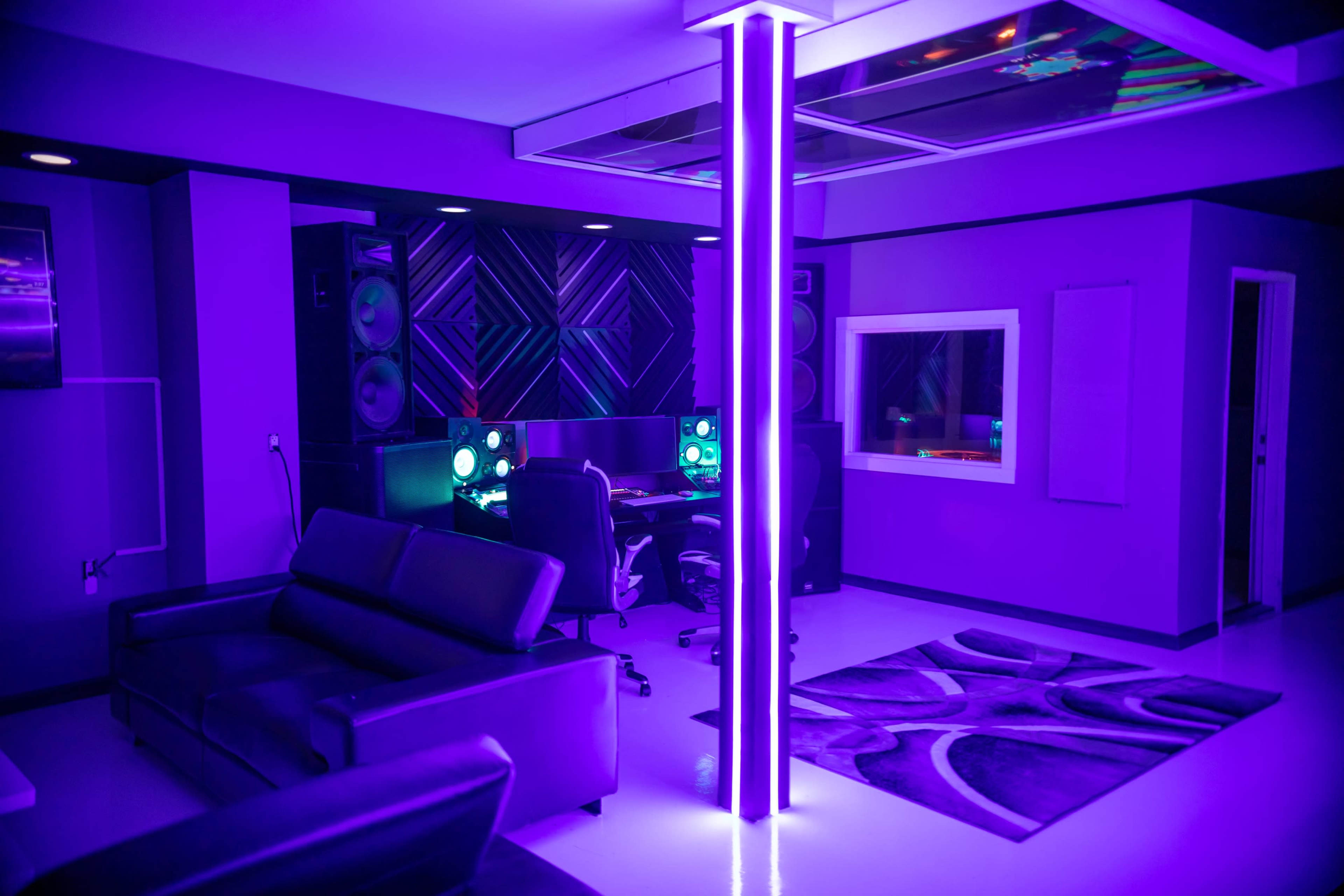 A modern recording studio with purple lighting, featuring two computer workstations, a black sofa, and large speakers.