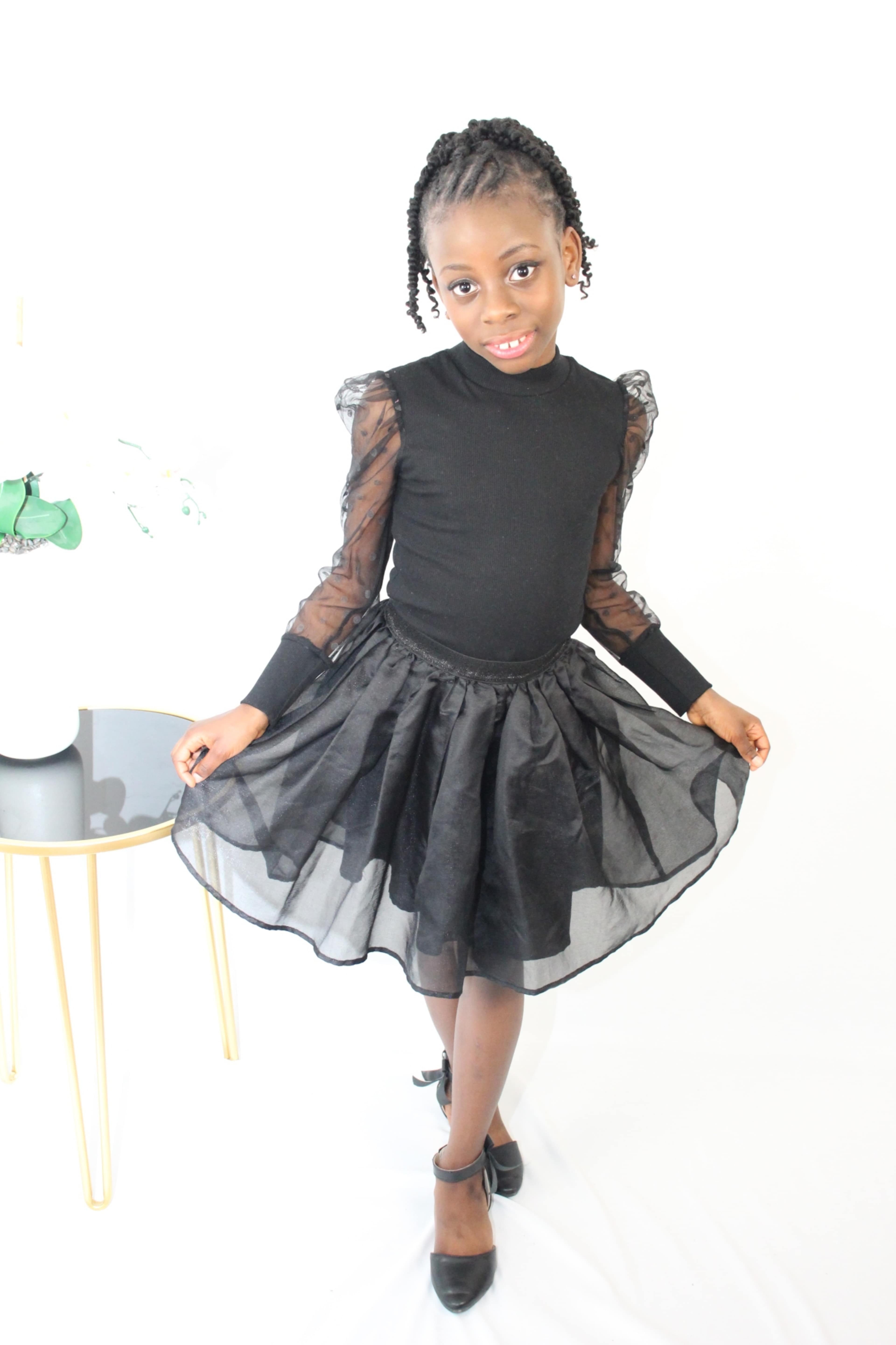 A girl in a black dress with sheer sleeves poses beside a small, round table.