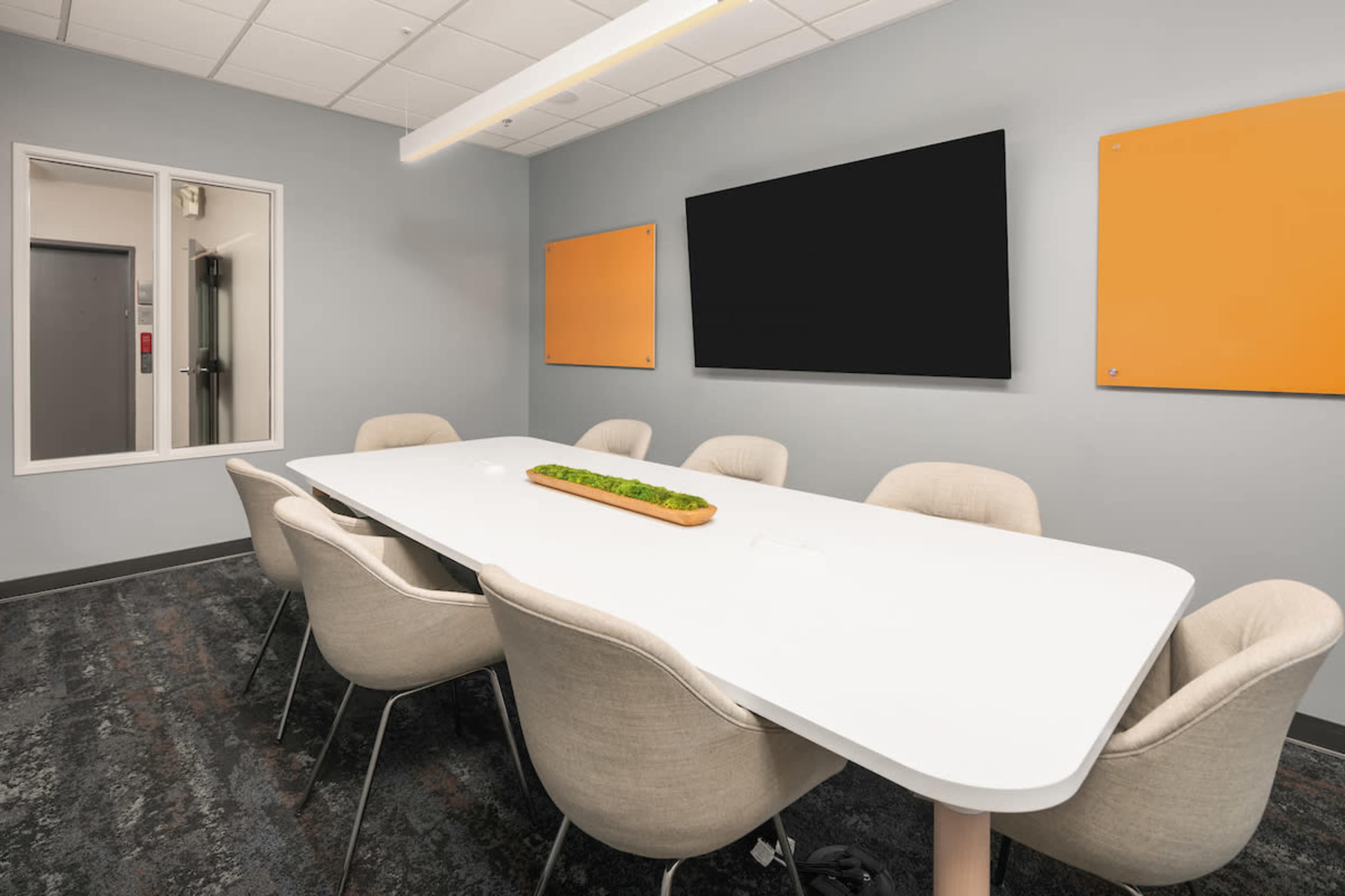 A modern conference room features a long white table surrounded by beige chairs, with a decorative planter in the center and two orange boards on the walls.
