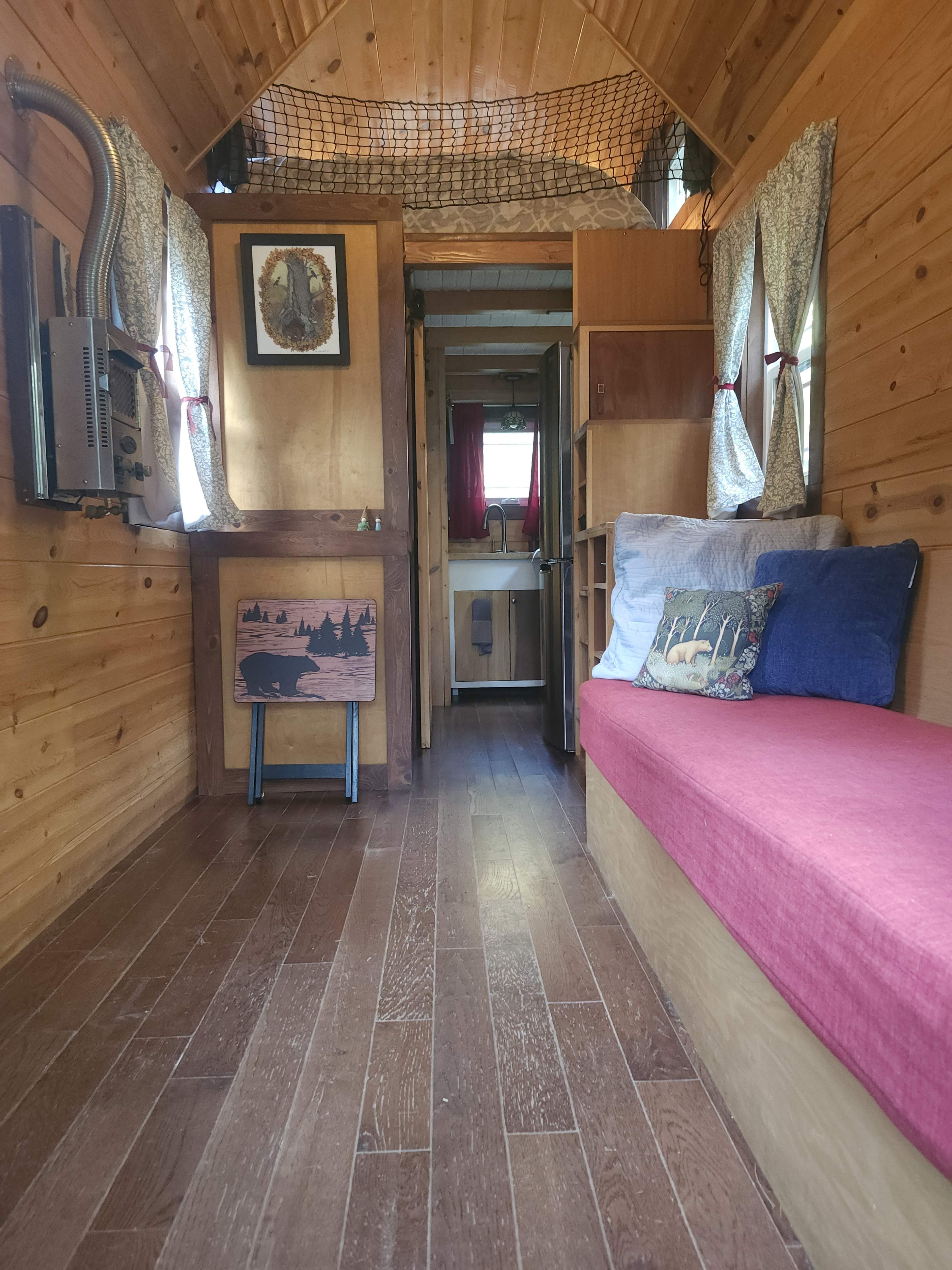 The interior of a wooden cabin features a red cushioned bench, a decorative wall hanging, and a view of a small kitchenette through a doorway.