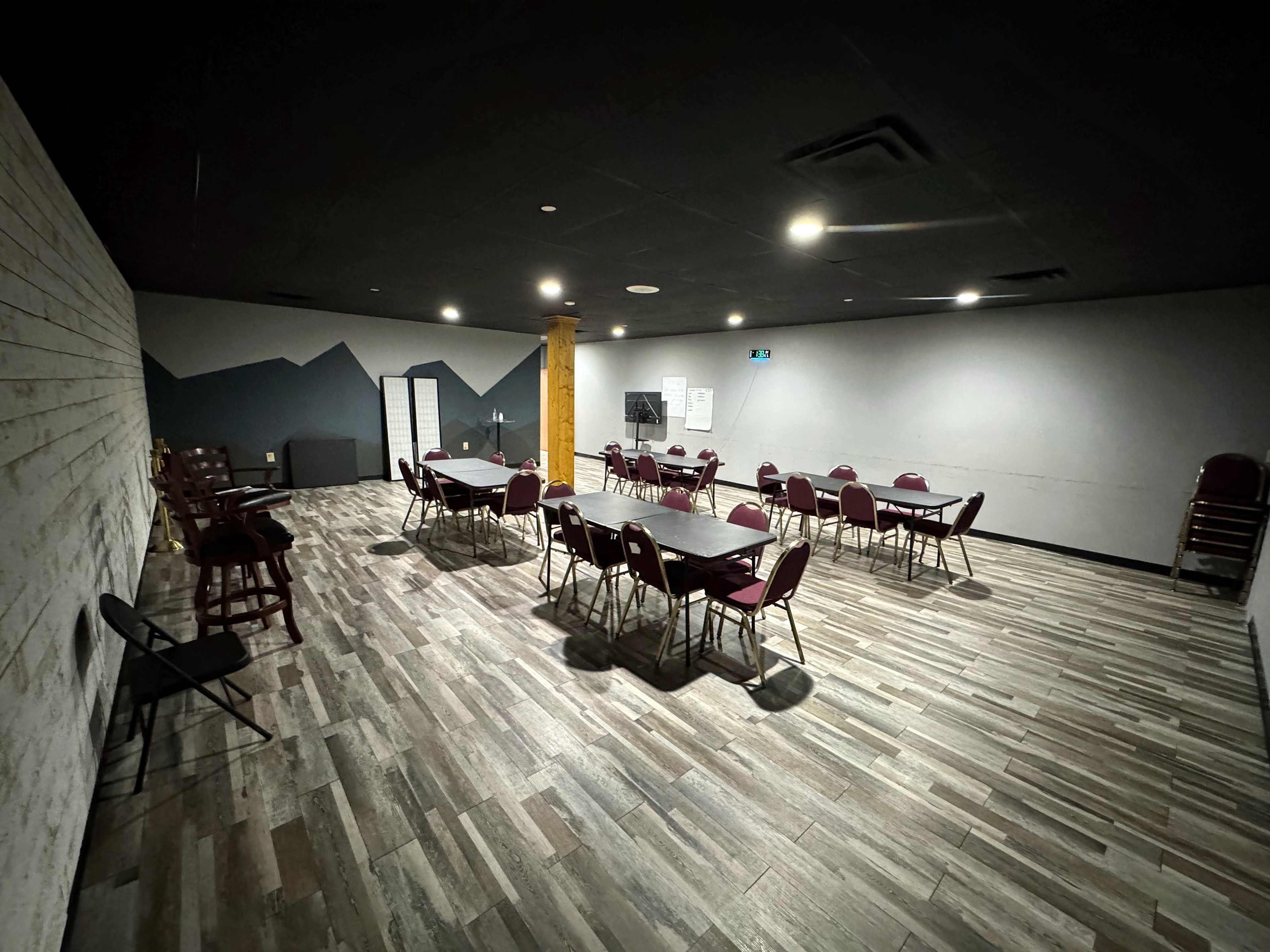 The image shows a spacious, empty meeting room with several tables and chairs arranged in a loose configuration, featuring a wall with a mountain mural in the background.