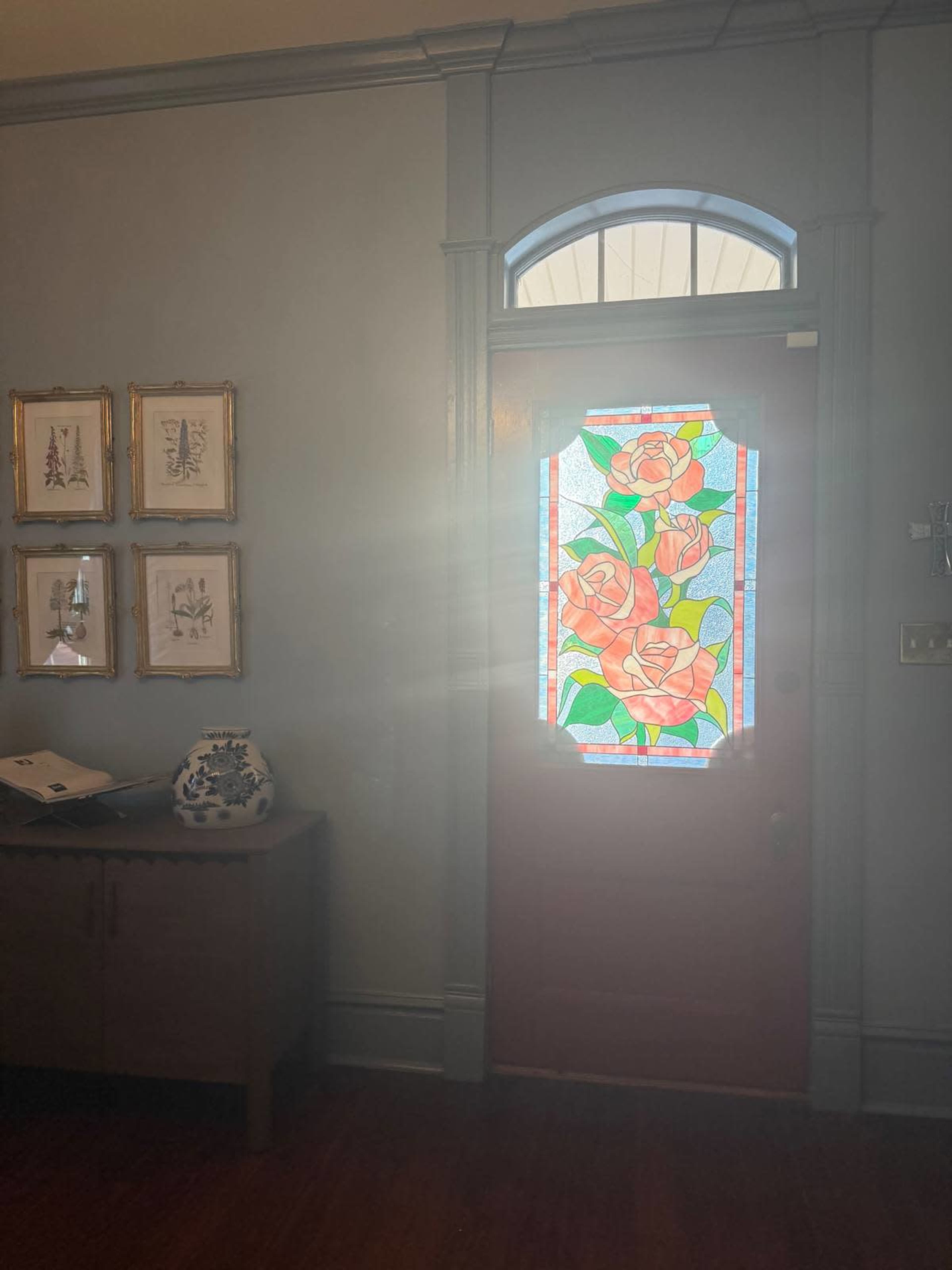 The image shows a wooden door with a stained glass panel depicting roses, illuminated by sunlight from outside.