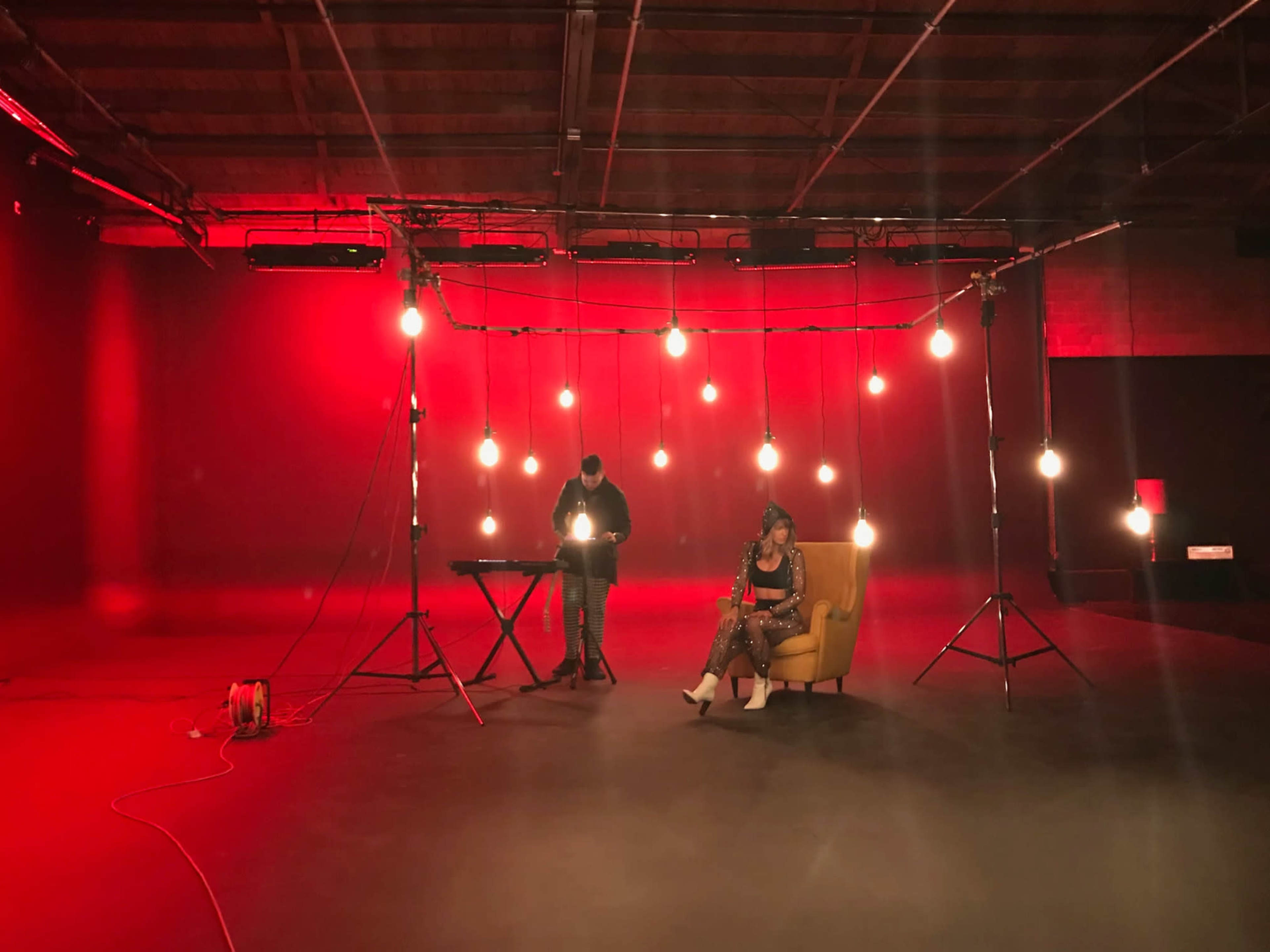A woman sits in a yellow chair under hanging lights while a man stands at a keyboard in a red-lit studio.