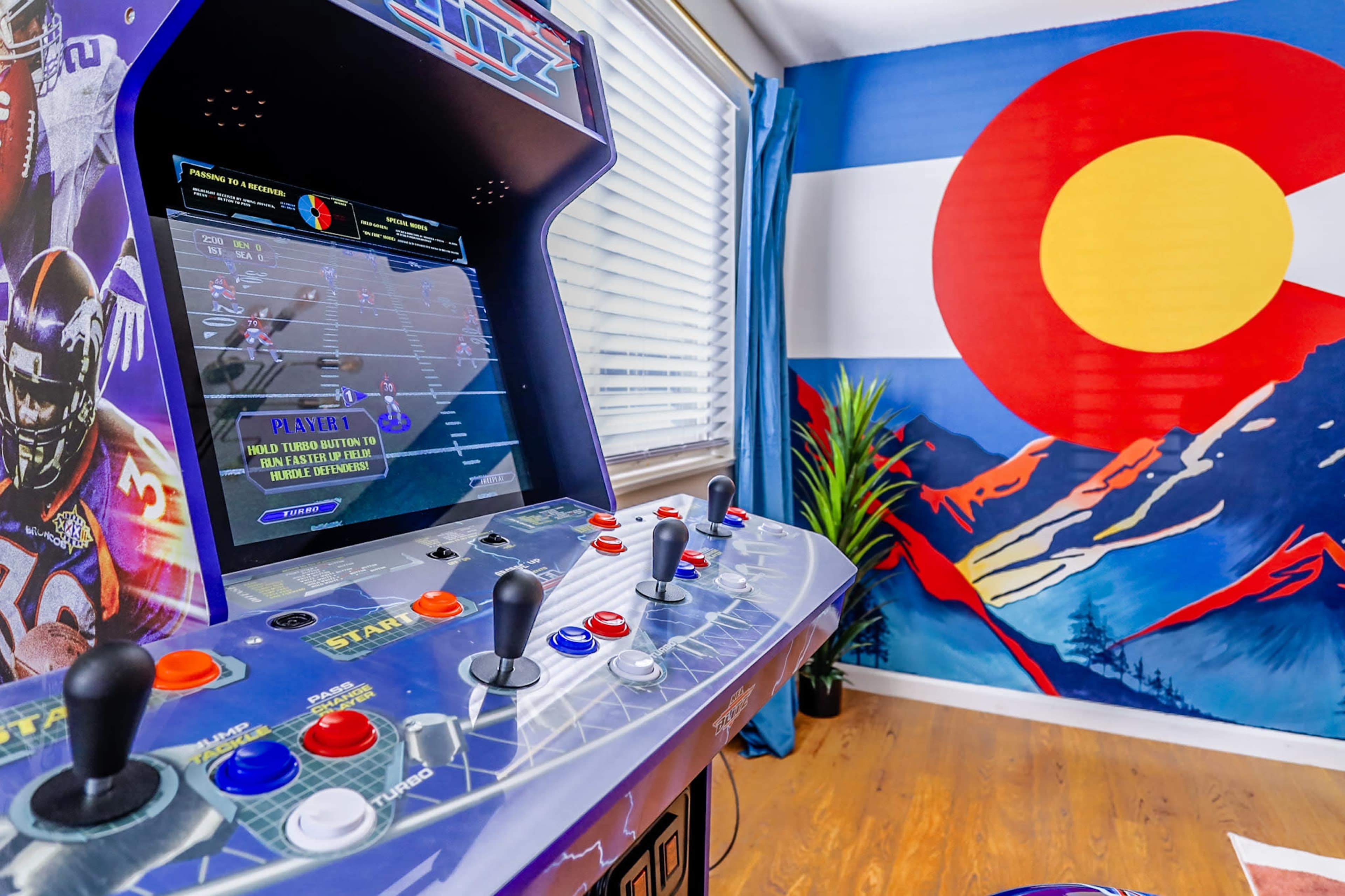 The image features a retro arcade game cabinet in a room with a large mural depicting the Colorado state flag and mountainous scenery.