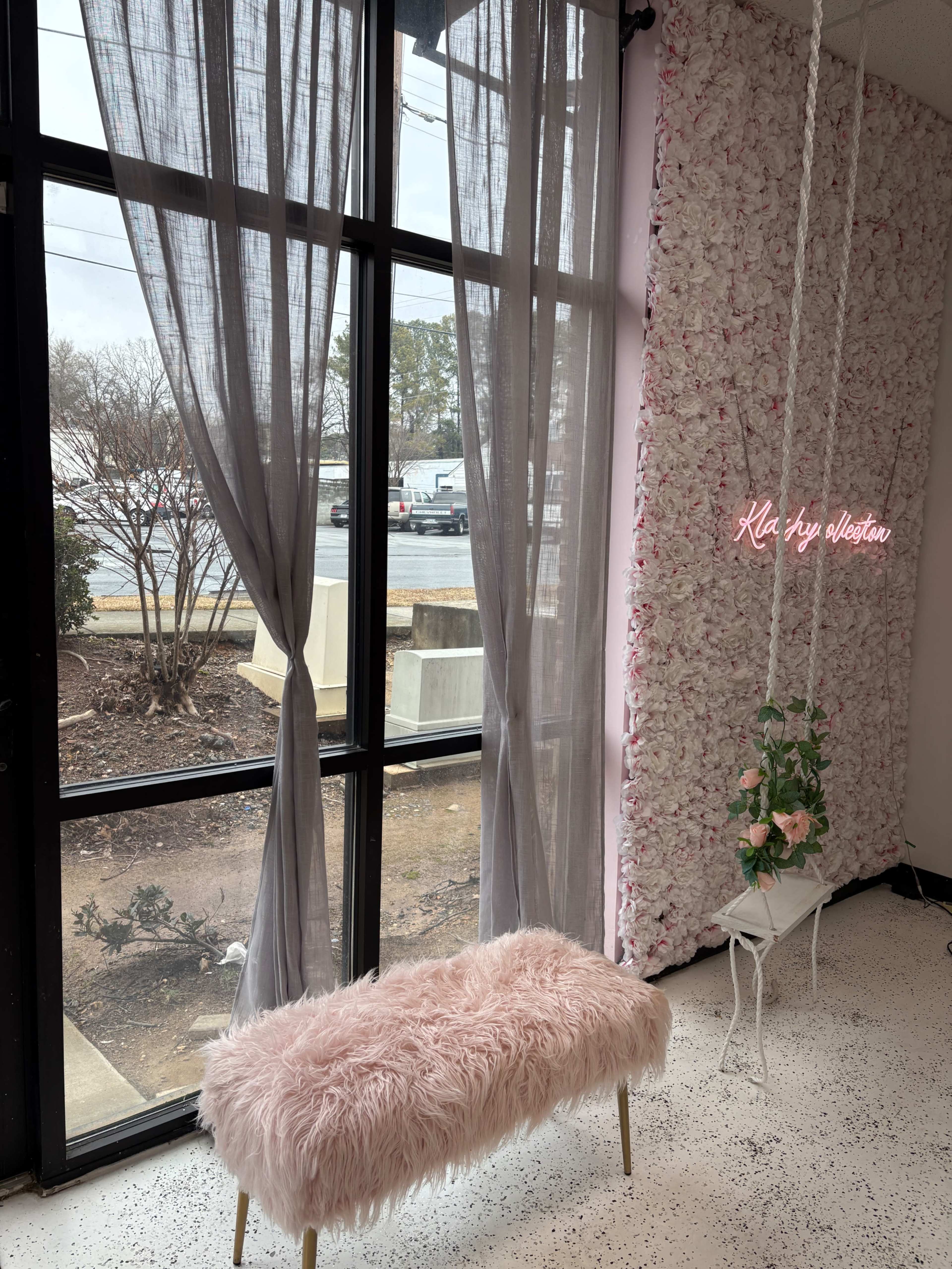 The image shows a bright interior with a pink fur bench, sheer gray curtains, and a floral wall, featuring a neon sign that reads "Kla Fly Aesthet."