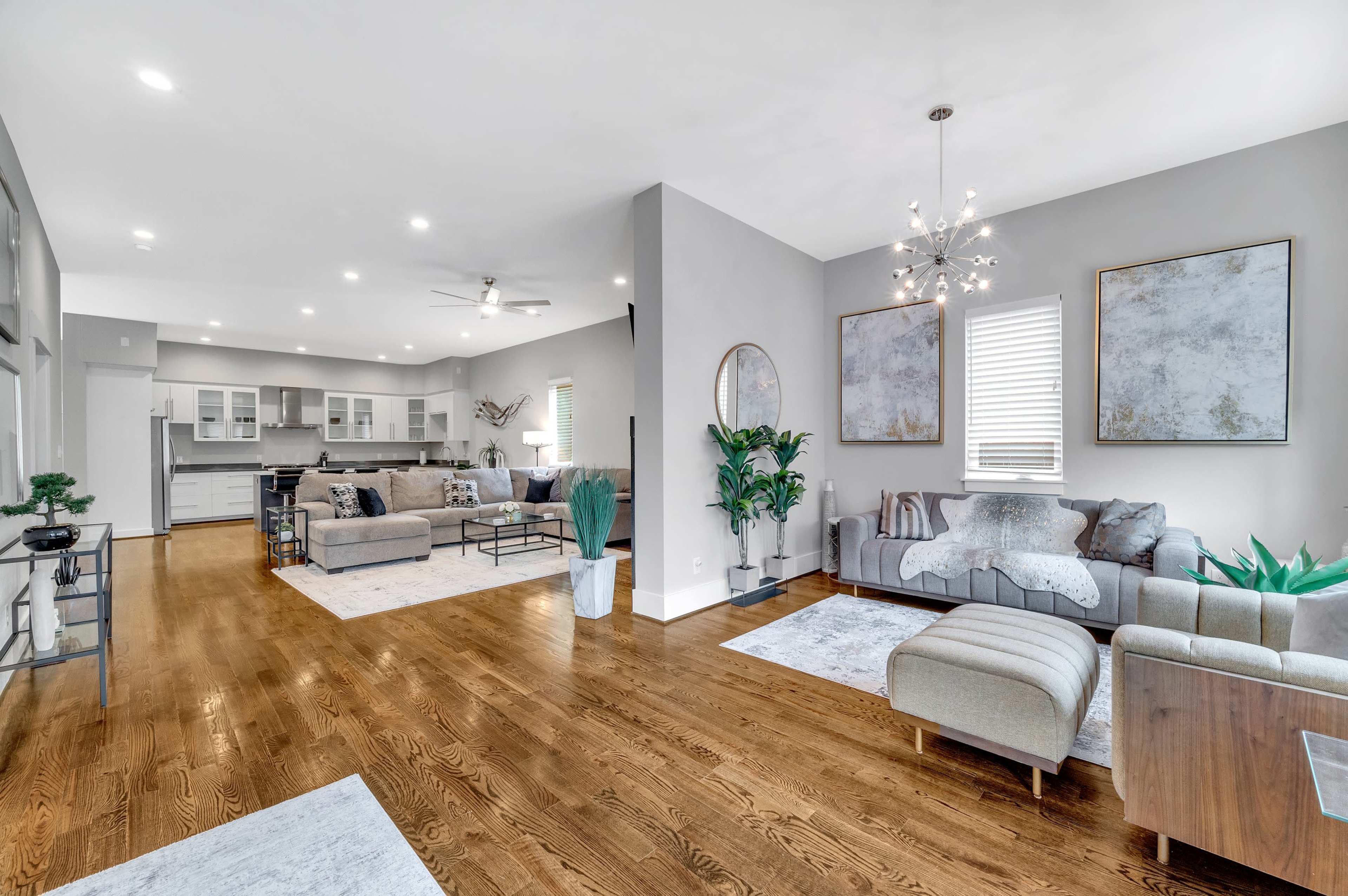A spacious living room features wood floors, modern furniture, and large windows, with an adjoining open kitchen.