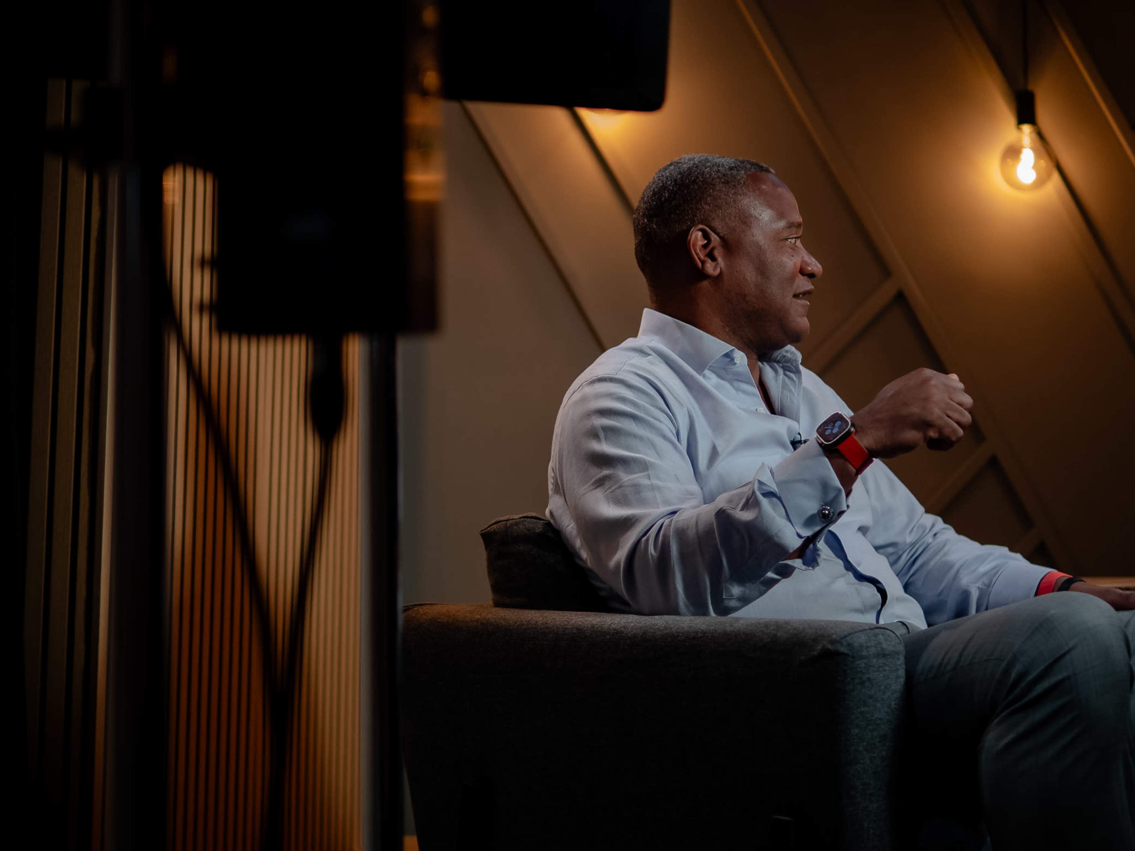A man seated on a gray couch engages in conversation, with warm lighting and textured walls in the background.