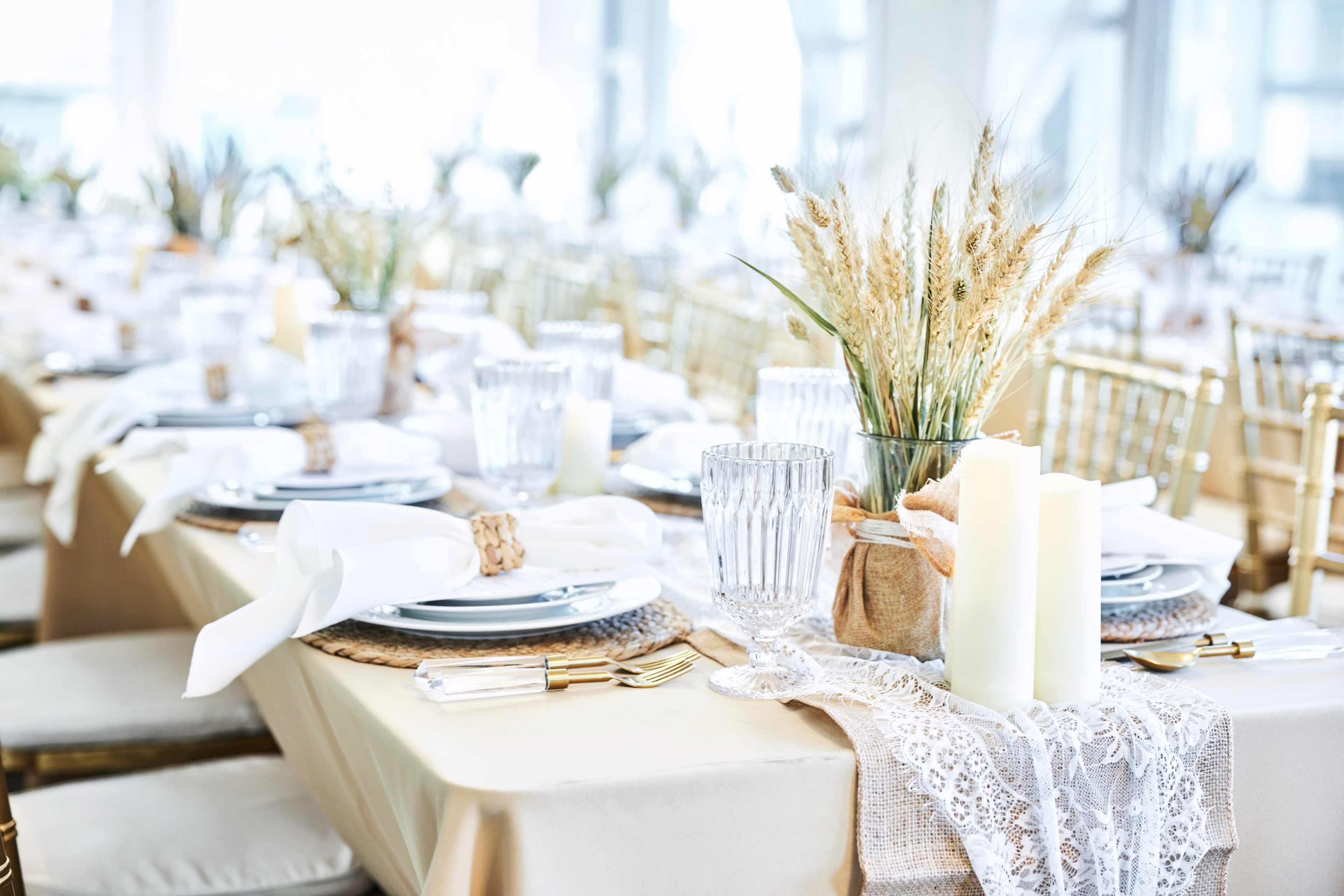 A long dining table is set with elegant tableware, decorative candles, and arrangements of dried wheat.