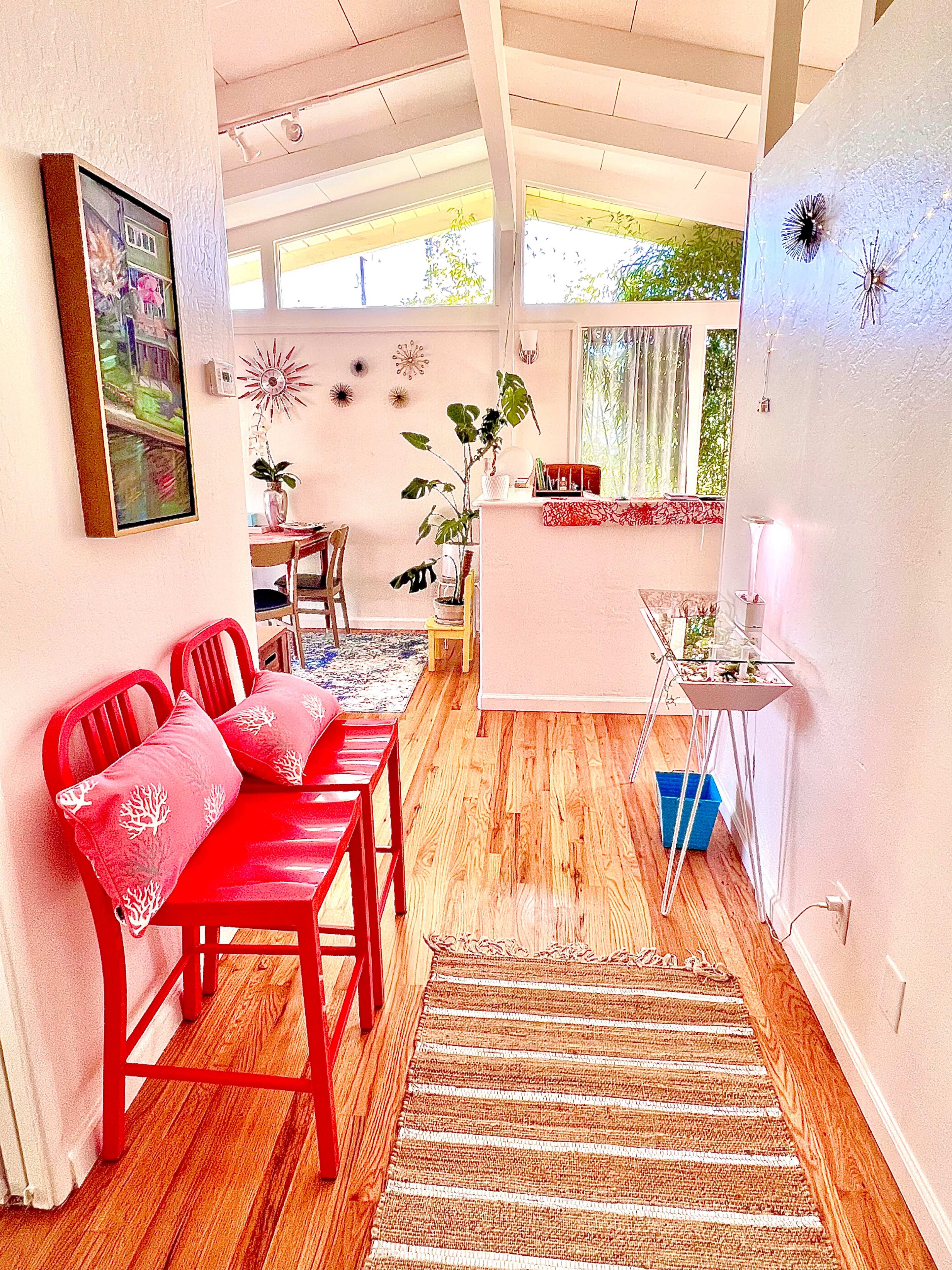 A bright hallway features red chairs with cushions, a small table adorned with decorative items, and a view of a dining area through large windows.