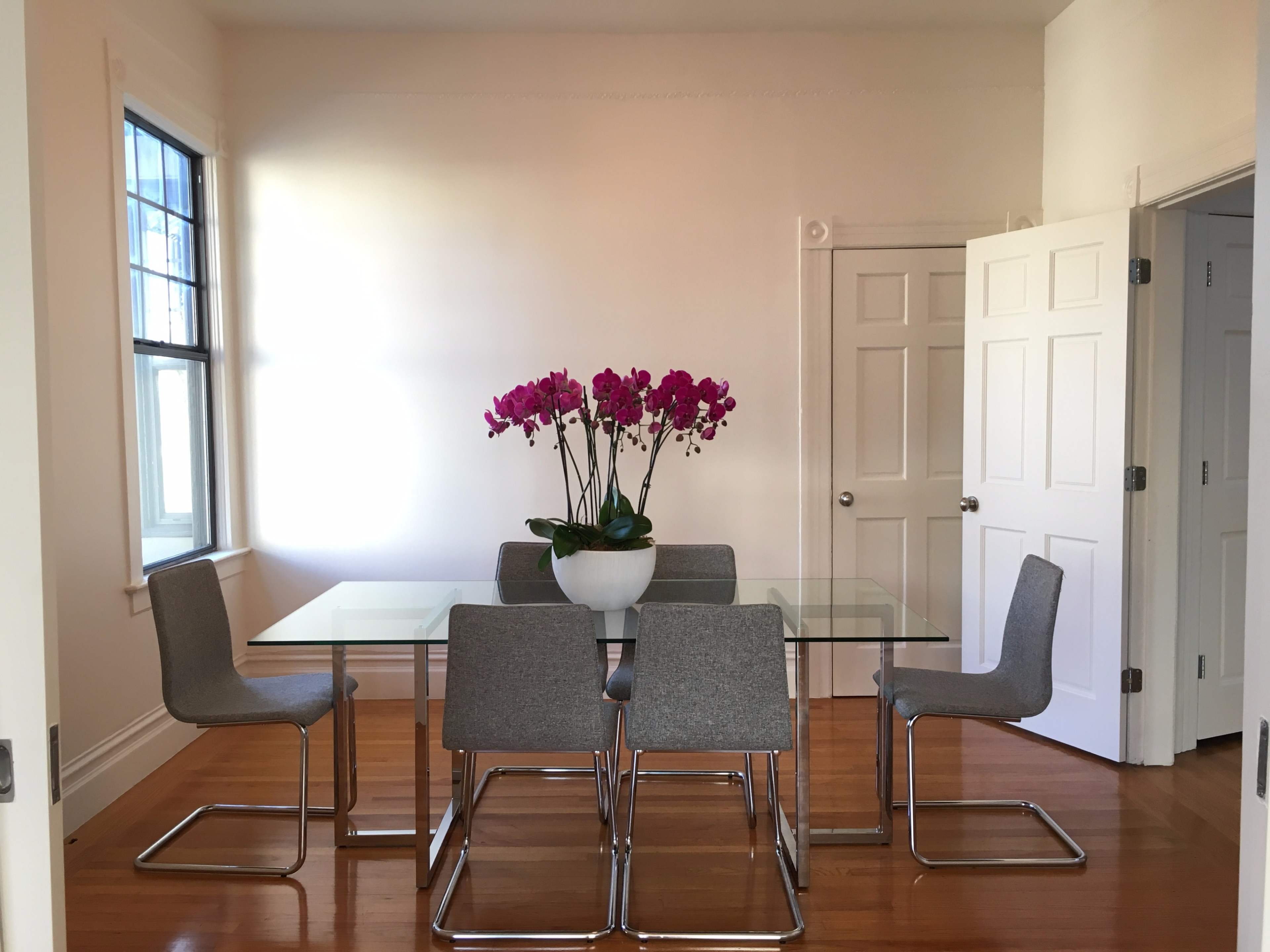 A glass dining table with five gray chairs and a potted orchid arrangement is positioned in a brightly lit room with white walls and two doors.