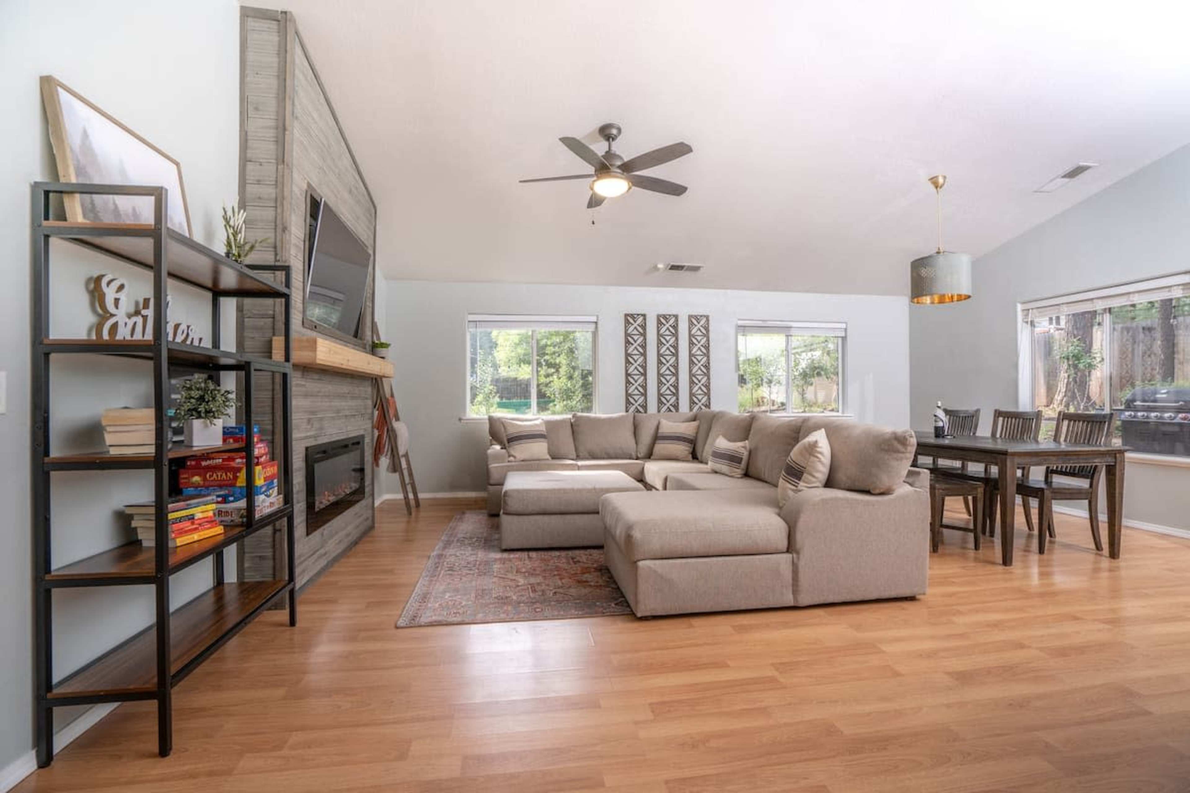 The image shows a spacious living area with a sectional sofa, a wall-mounted television above a fireplace, a dining table, and large windows letting in natural light.
