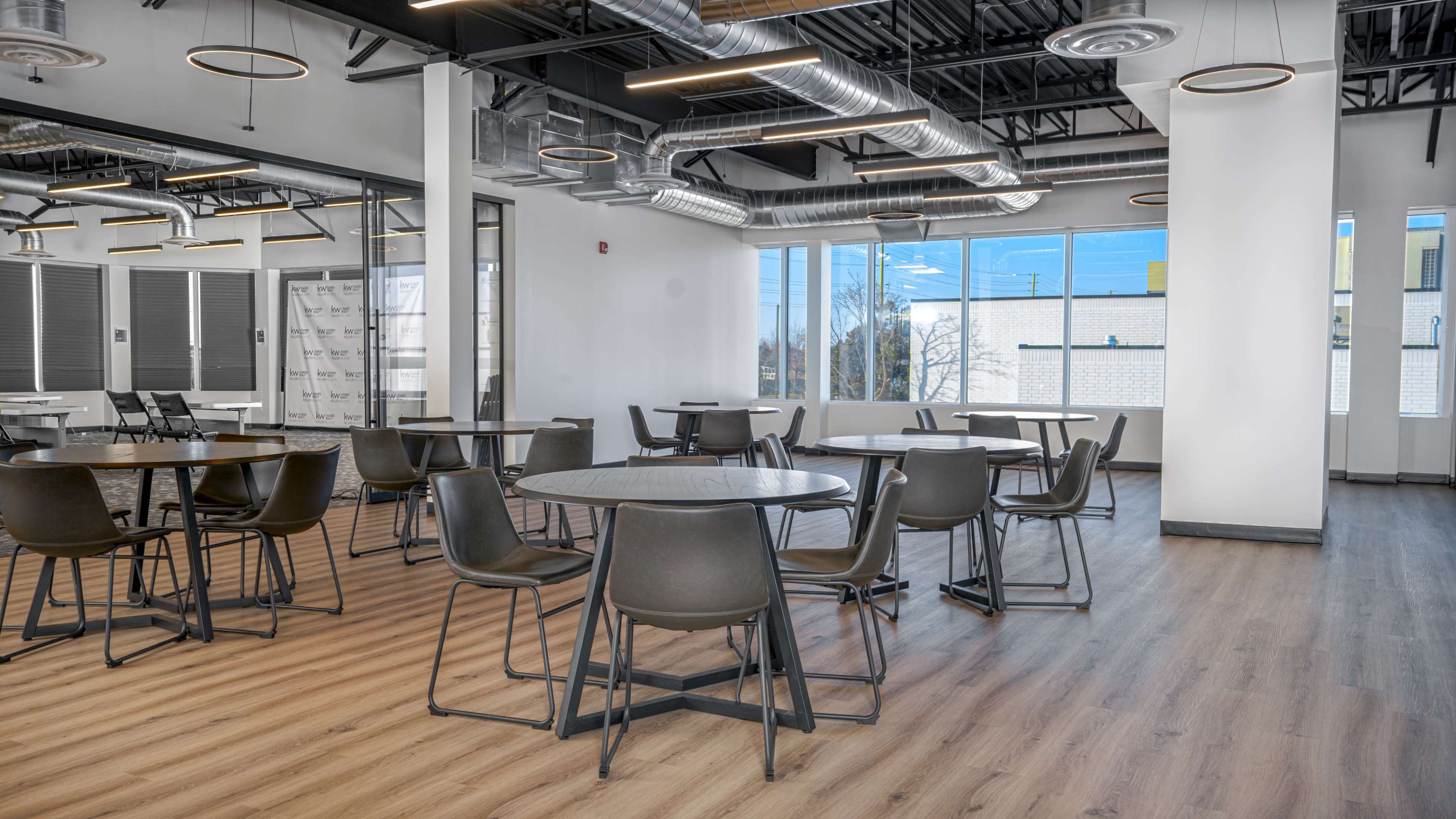 A modern, open workspace with several round tables and chairs arranged on a wood floor, surrounded by large windows and sleek ceiling fixtures.