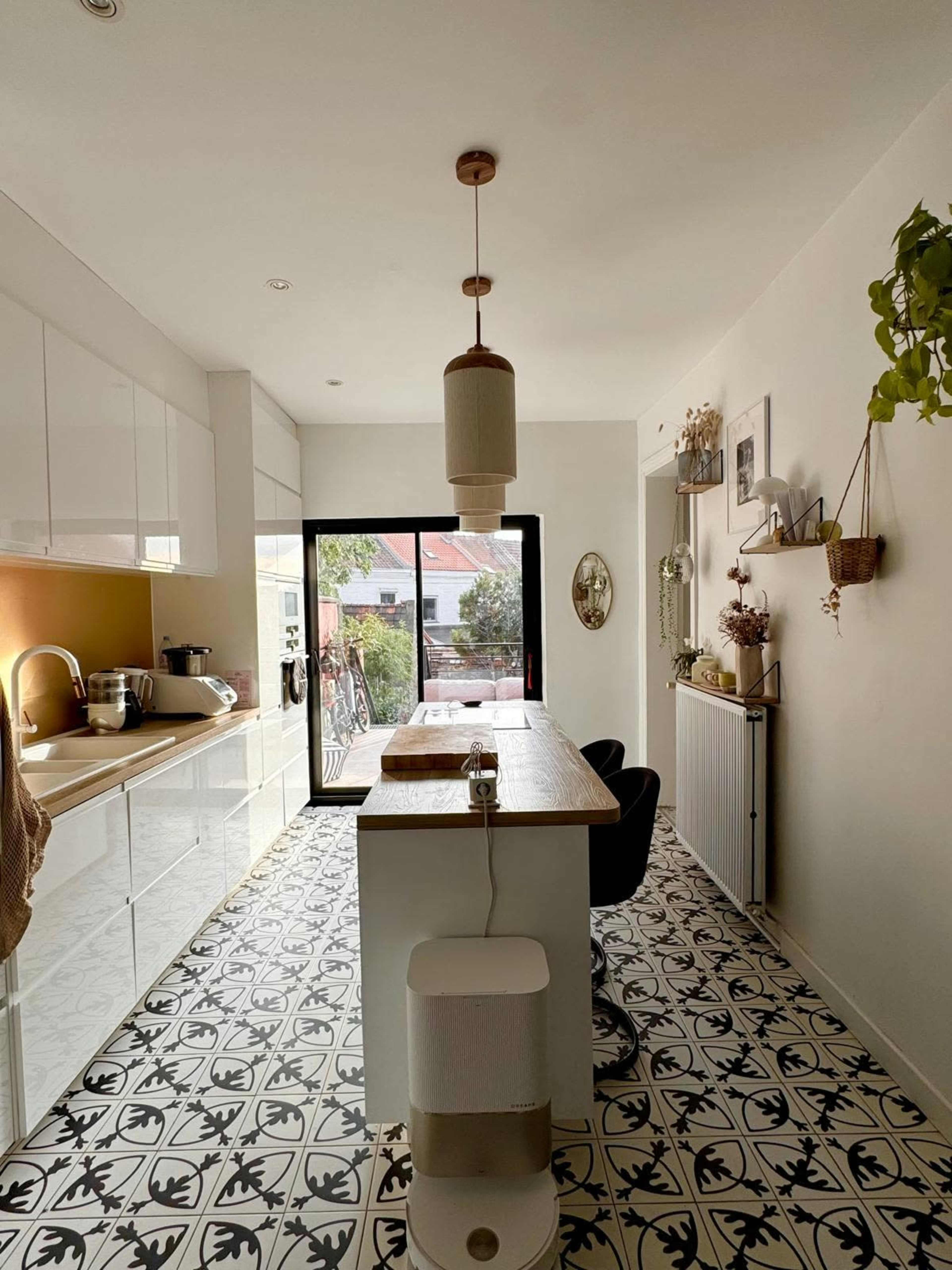 The image shows a modern kitchen with white cabinets, a wooden dining table, and patterned tile flooring, illuminated by natural light coming through a large glass door.