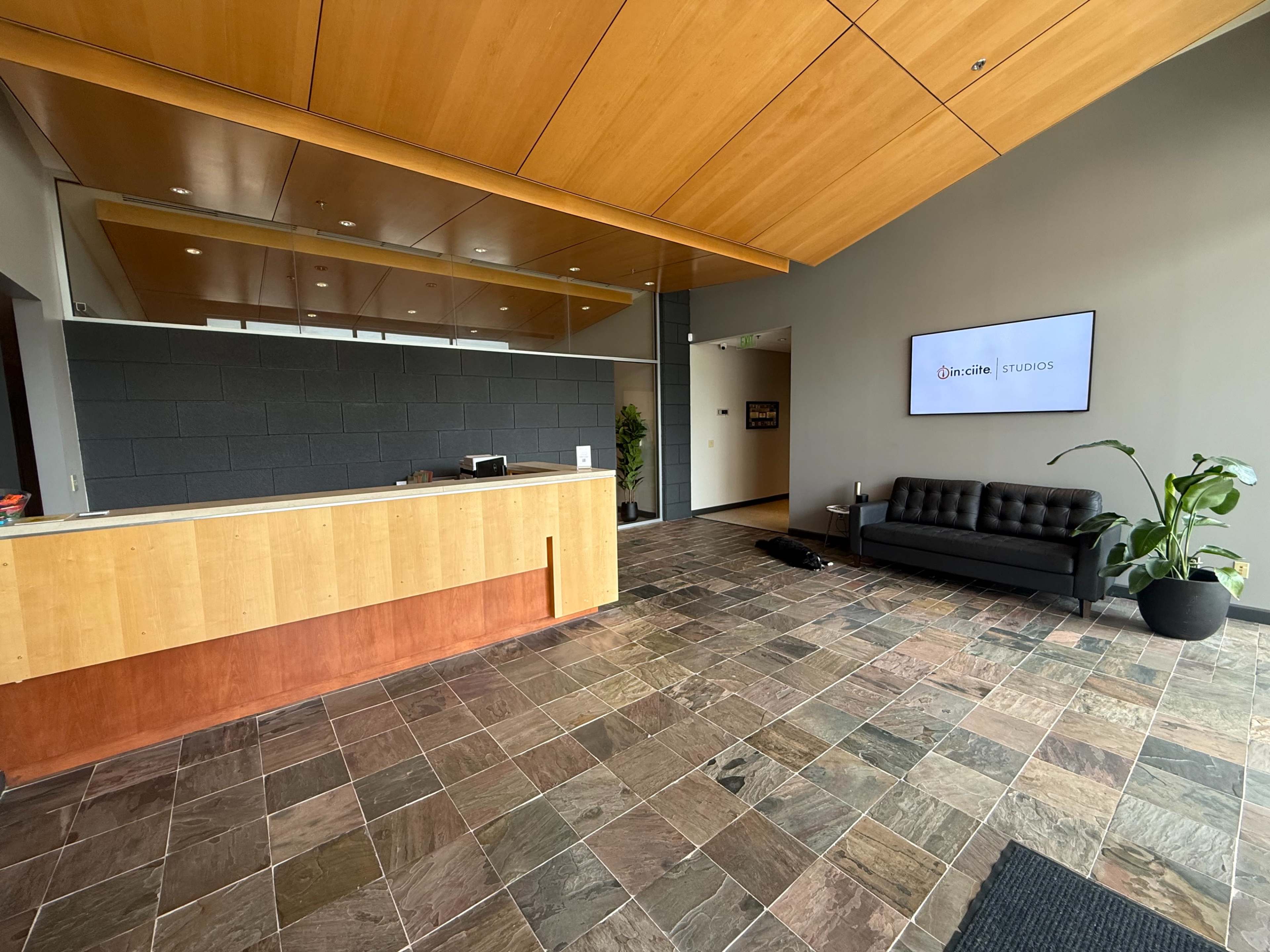 The image shows a modern reception area featuring a wooden front desk, a black couch, and a large plant, with slate tile flooring and a digital screen displaying "Quincy Studios."