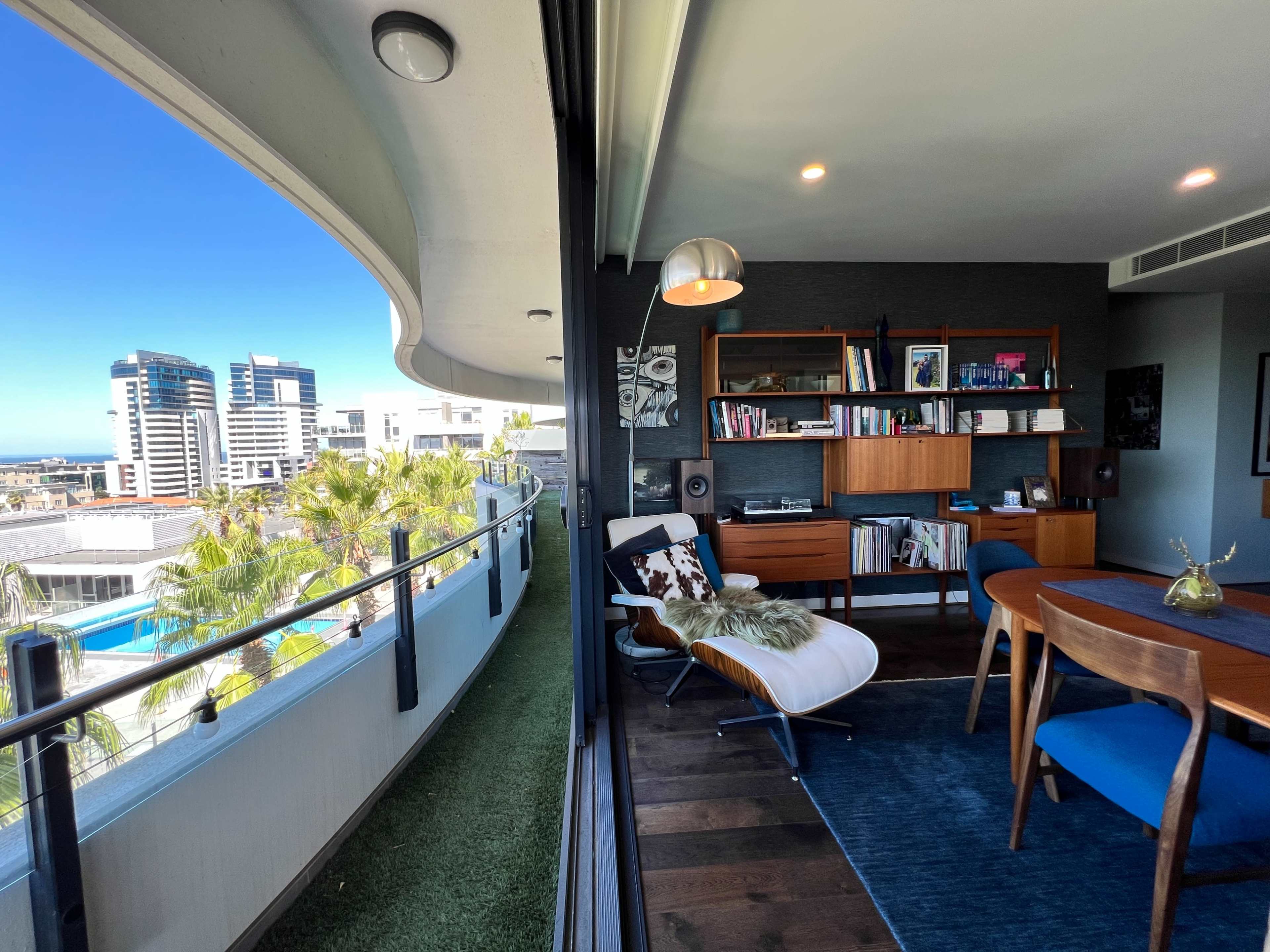 The image shows a modern balcony overlooking a city skyline, with a seating area and a well-furnished interior space that includes a bookcase and dining table.