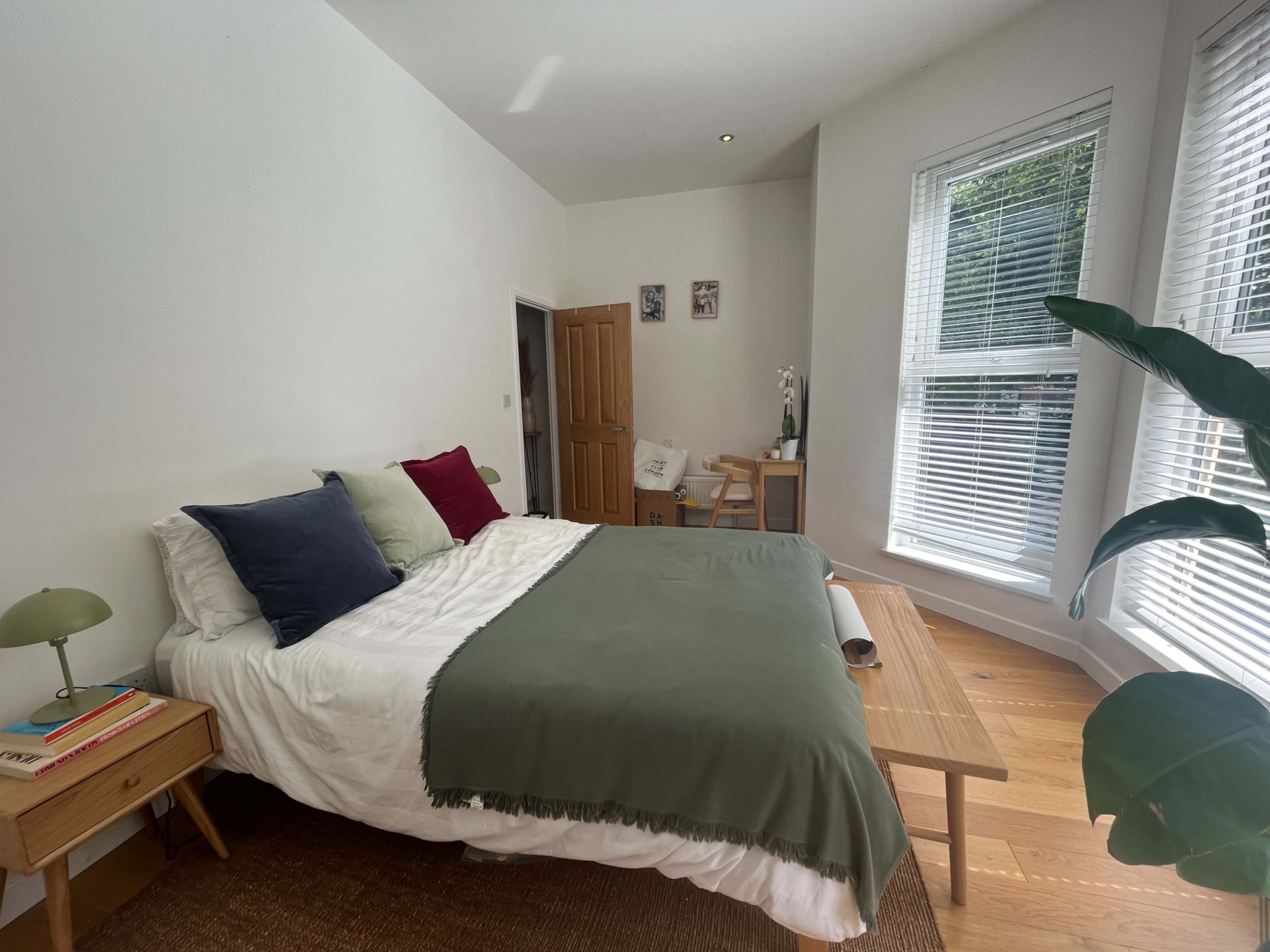 A bright bedroom features a large bed with a green throw, a bedside table with a lamp, and a window with blinds letting in natural light.