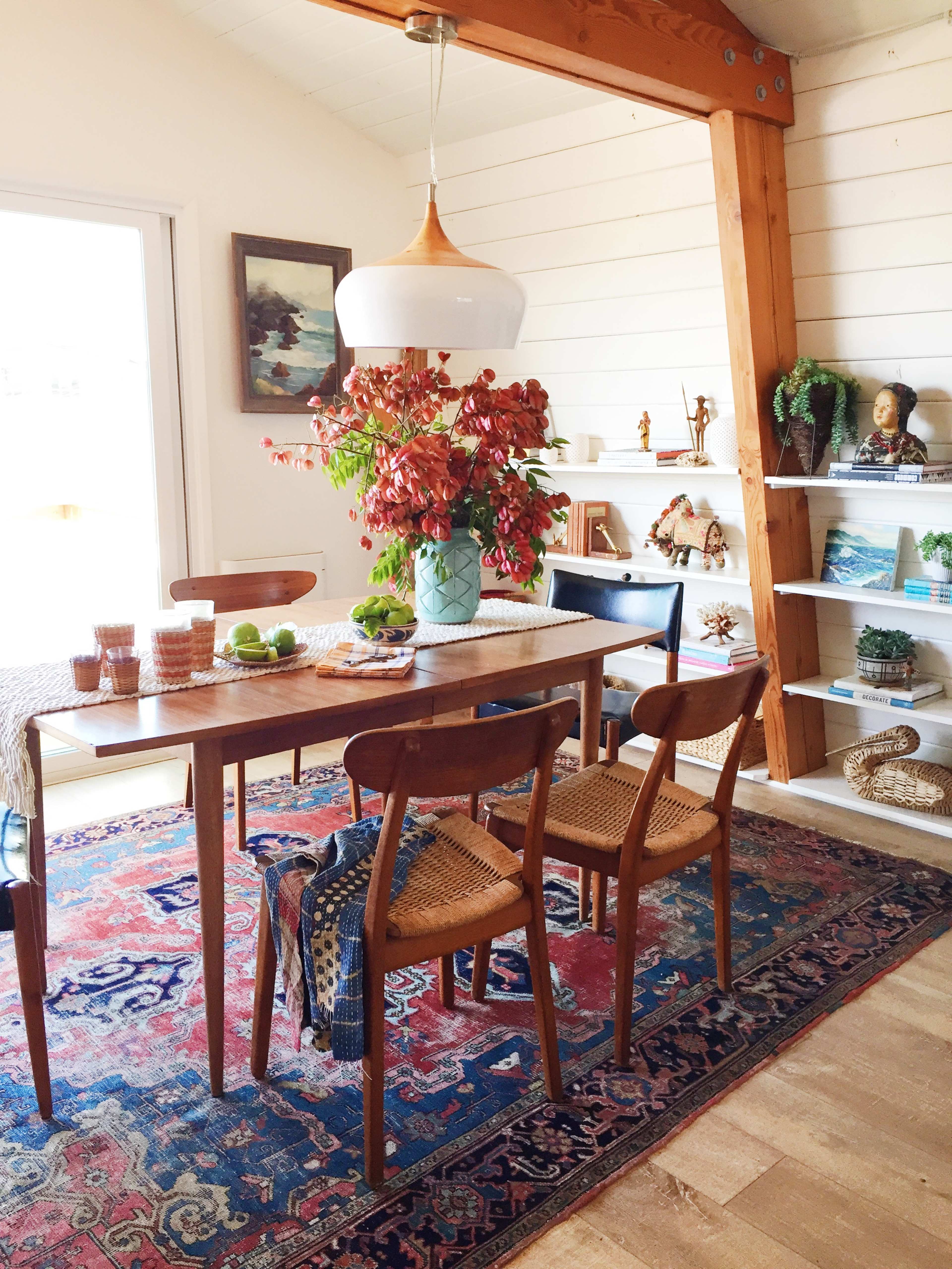 A dining area features a wooden table with four chairs, a large vase of flowers as the centerpiece, and shelves displaying various decorative items.