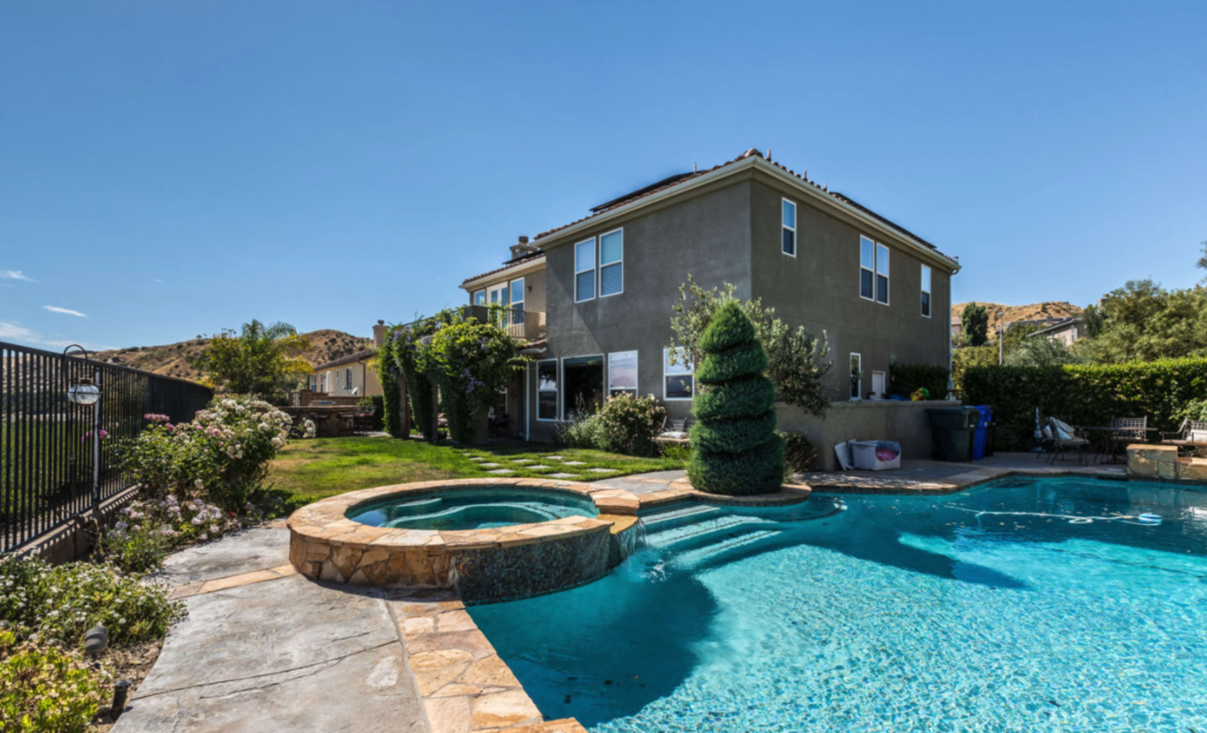 A two-story house with a fenced backyard features a swimming pool and a spa surrounded by landscaping and hills in the background.