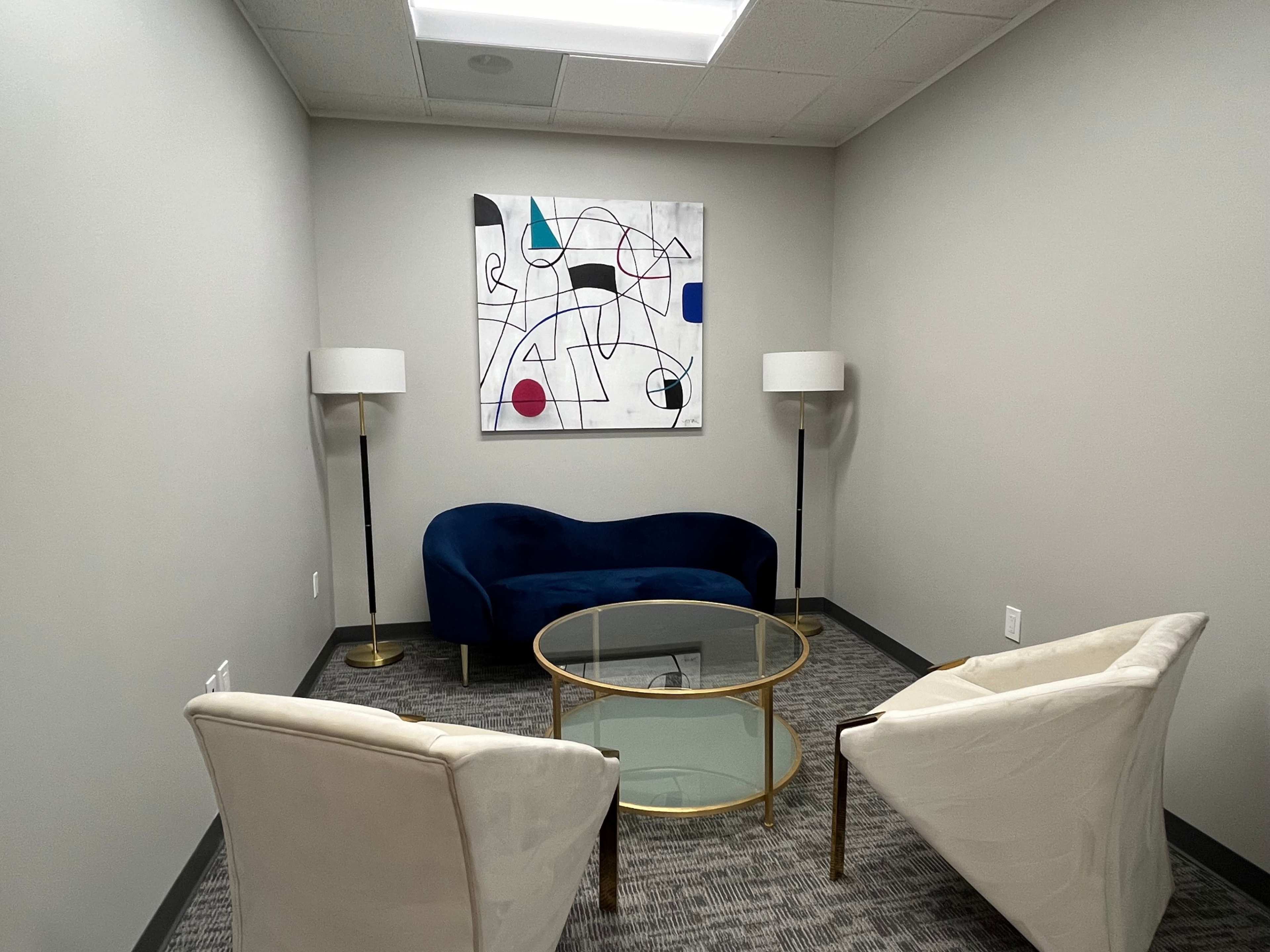 A small meeting room features a blue couch, two white chairs, a glass coffee table, and a colorful abstract painting on the wall.