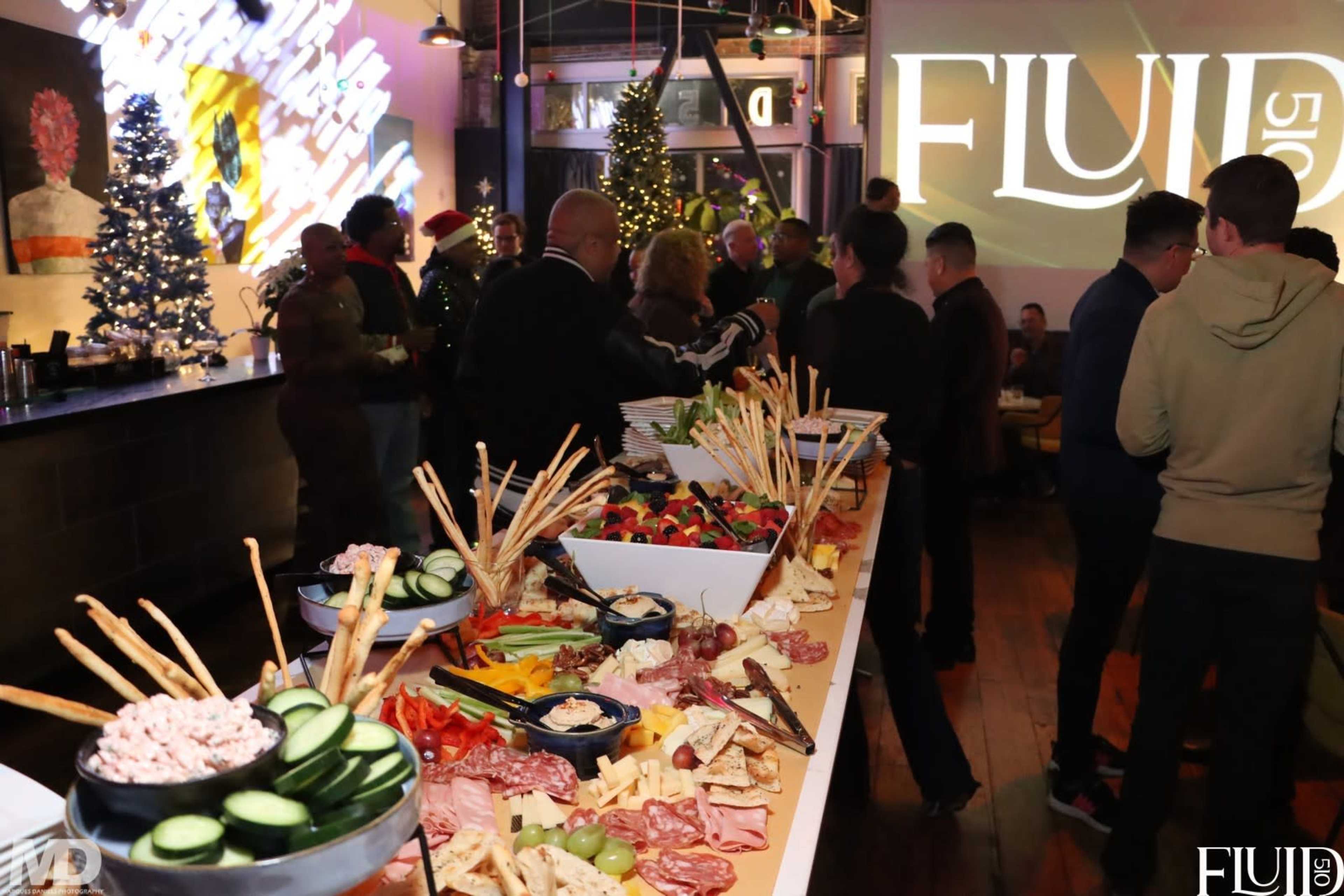 A long buffet table is filled with a variety of foods, including fruits, cheeses, meats, and snacks, while guests socialize in a festive setting decorated with Christmas trees.