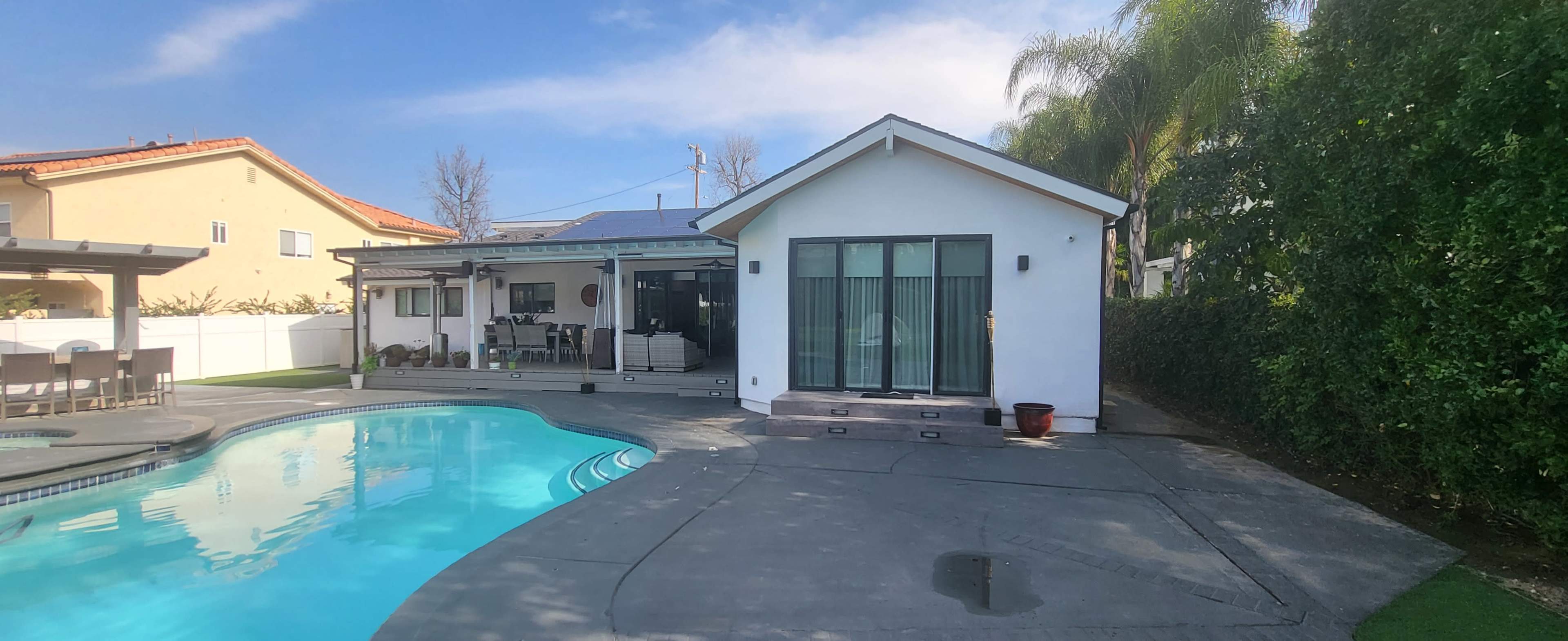 A modern house with large windows is situated beside a swimming pool, surrounded by palm trees and greenery.