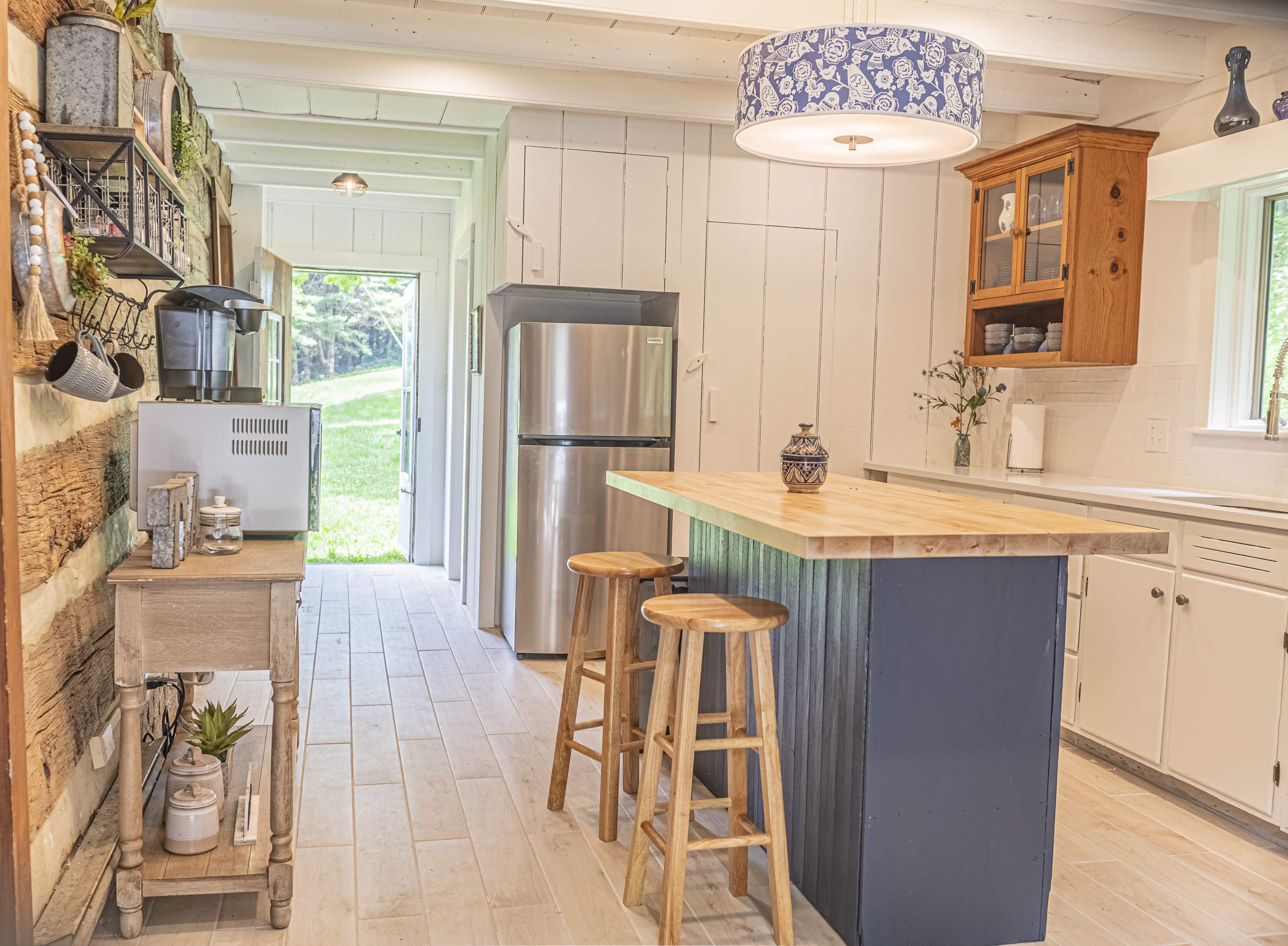 A modern kitchen features wooden cabinetry, a central island with stools, and a large window that opens to a grassy outdoor area.