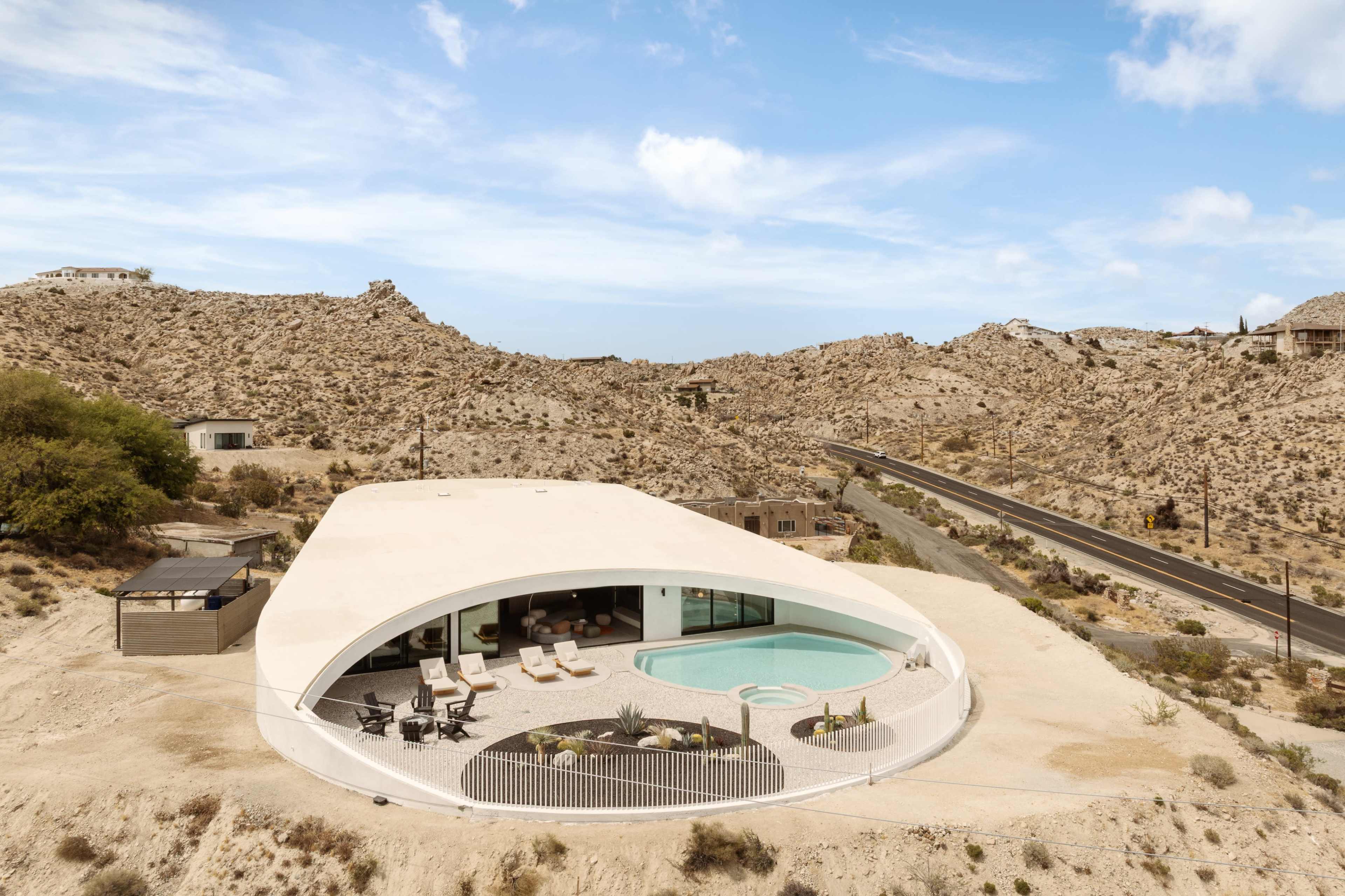A modern, dome-shaped house with a pool sits on a rocky landscape, surrounded by sparse vegetation and a road in the distance.