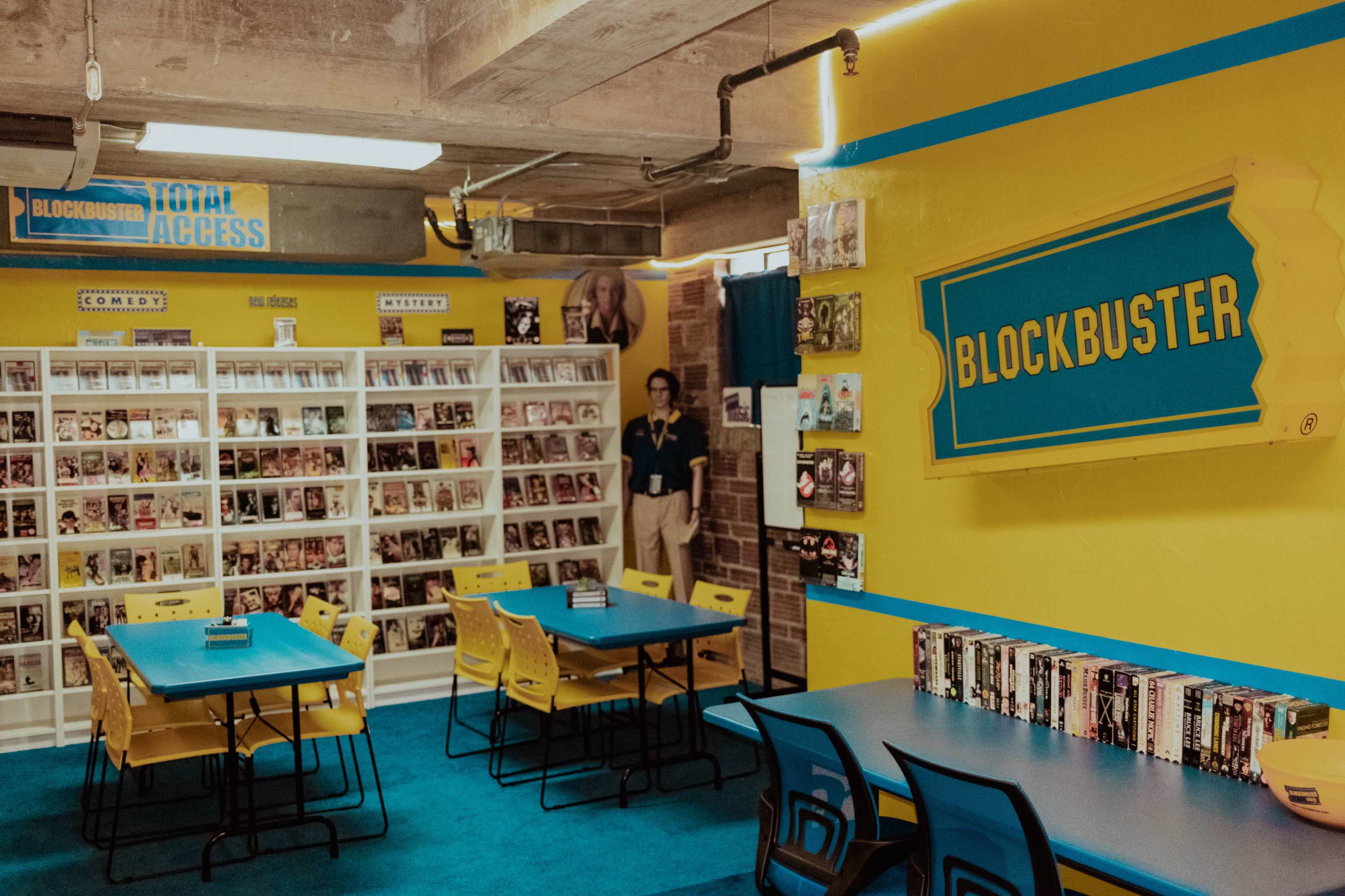 The image shows a brightly colored room resembling an old-fashioned video rental store, featuring shelves filled with movies and several tables with chairs arranged for seating.
