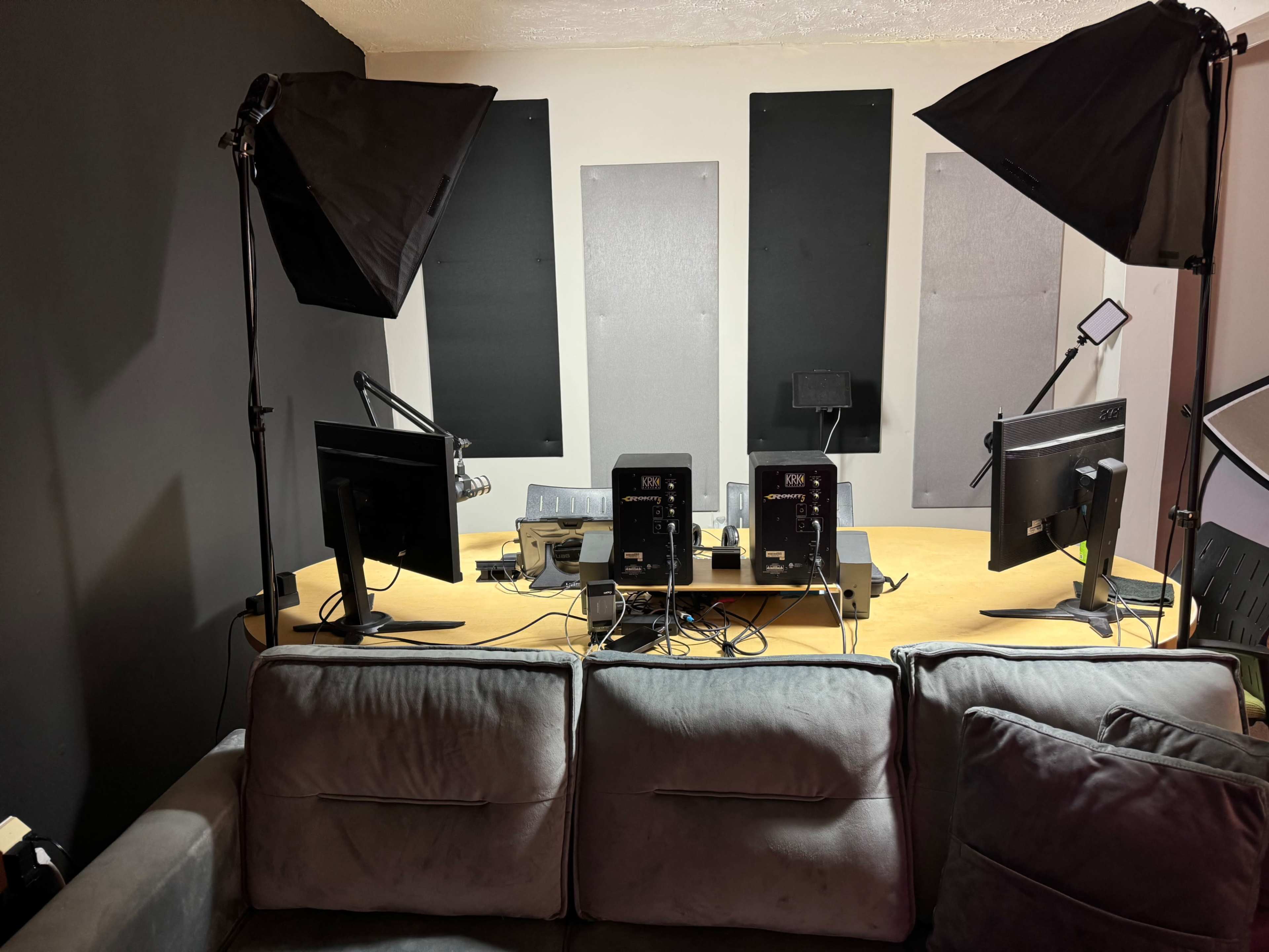 A workspace featuring a couch in the foreground and a desk with two computer monitors, several microphones, and lighting equipment in a dimly lit room.