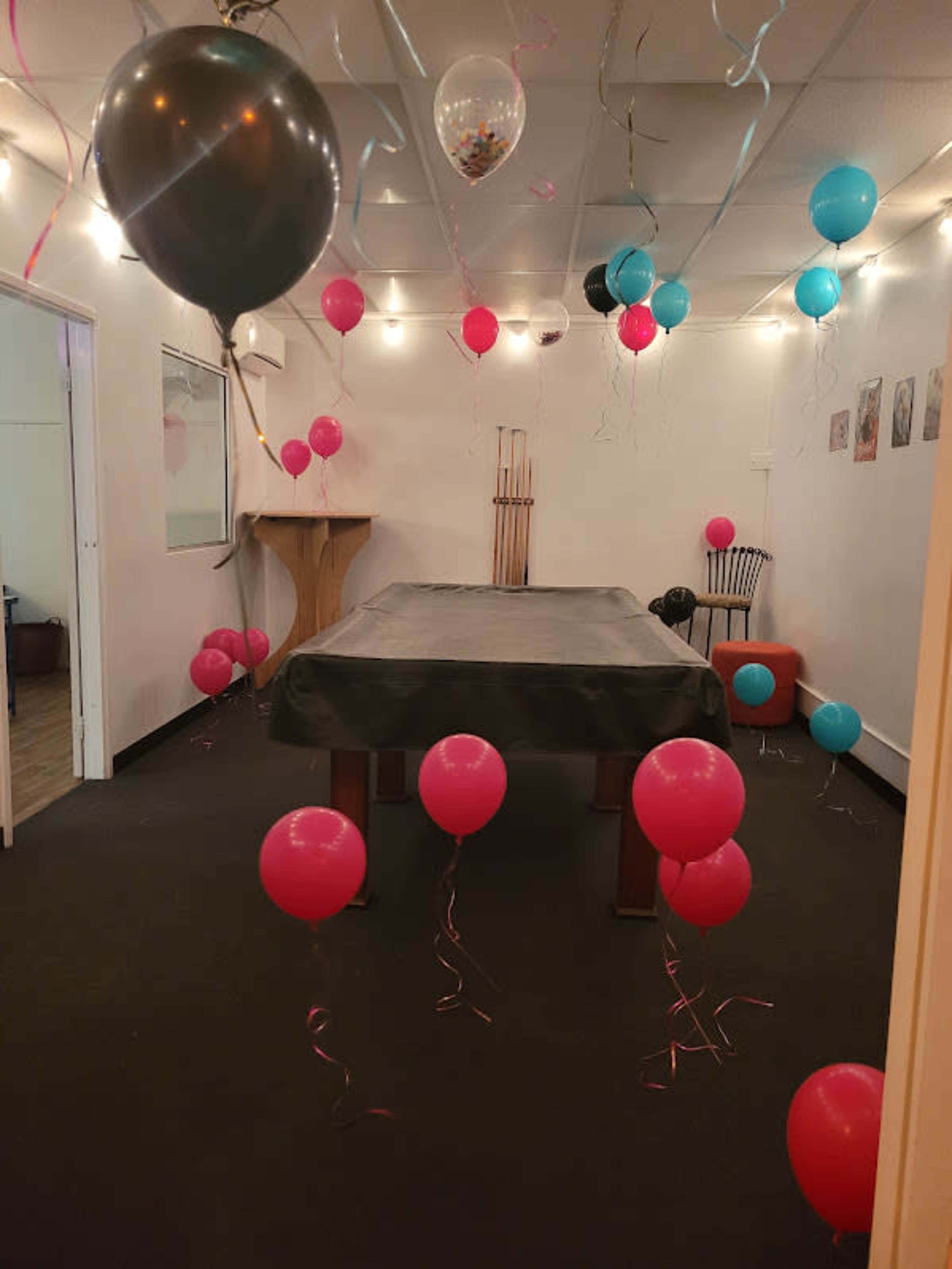 The room features a pool table surrounded by pink and blue balloons, with additional balloons floating from the ceiling and string decorations.