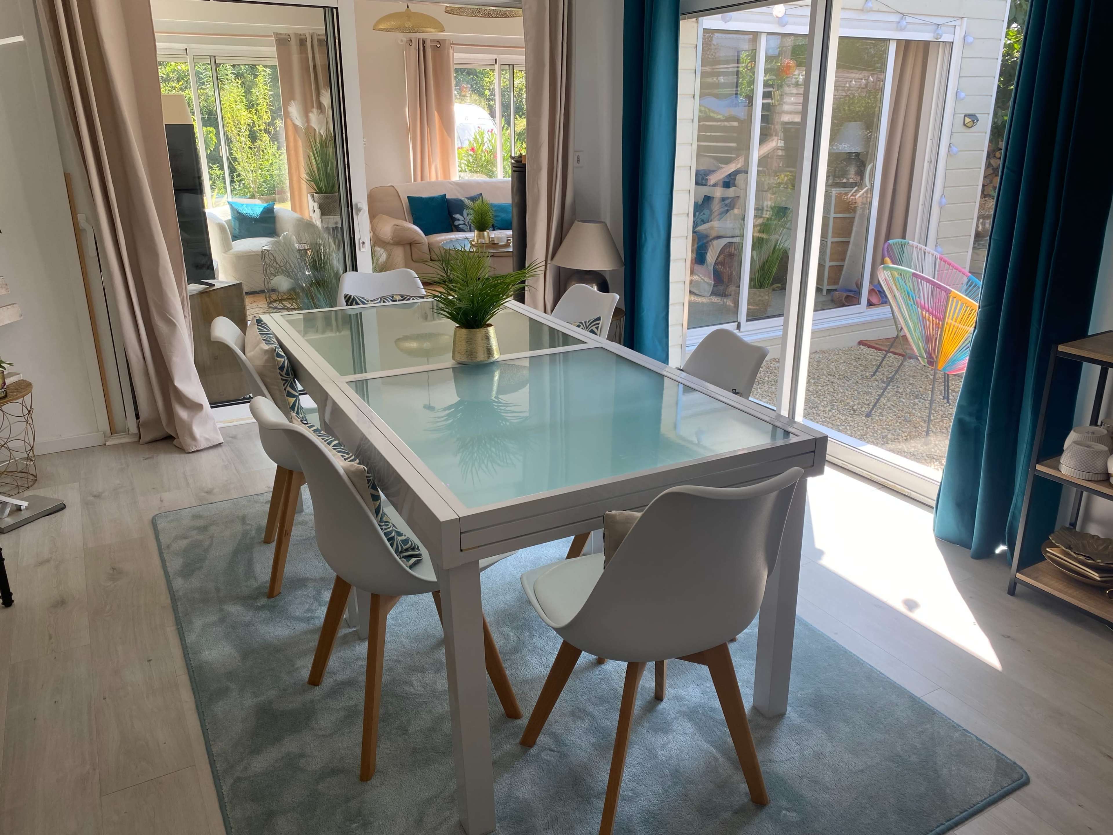 A modern dining area features a glass-top table surrounded by white chairs, with natural light streaming in from large windows.