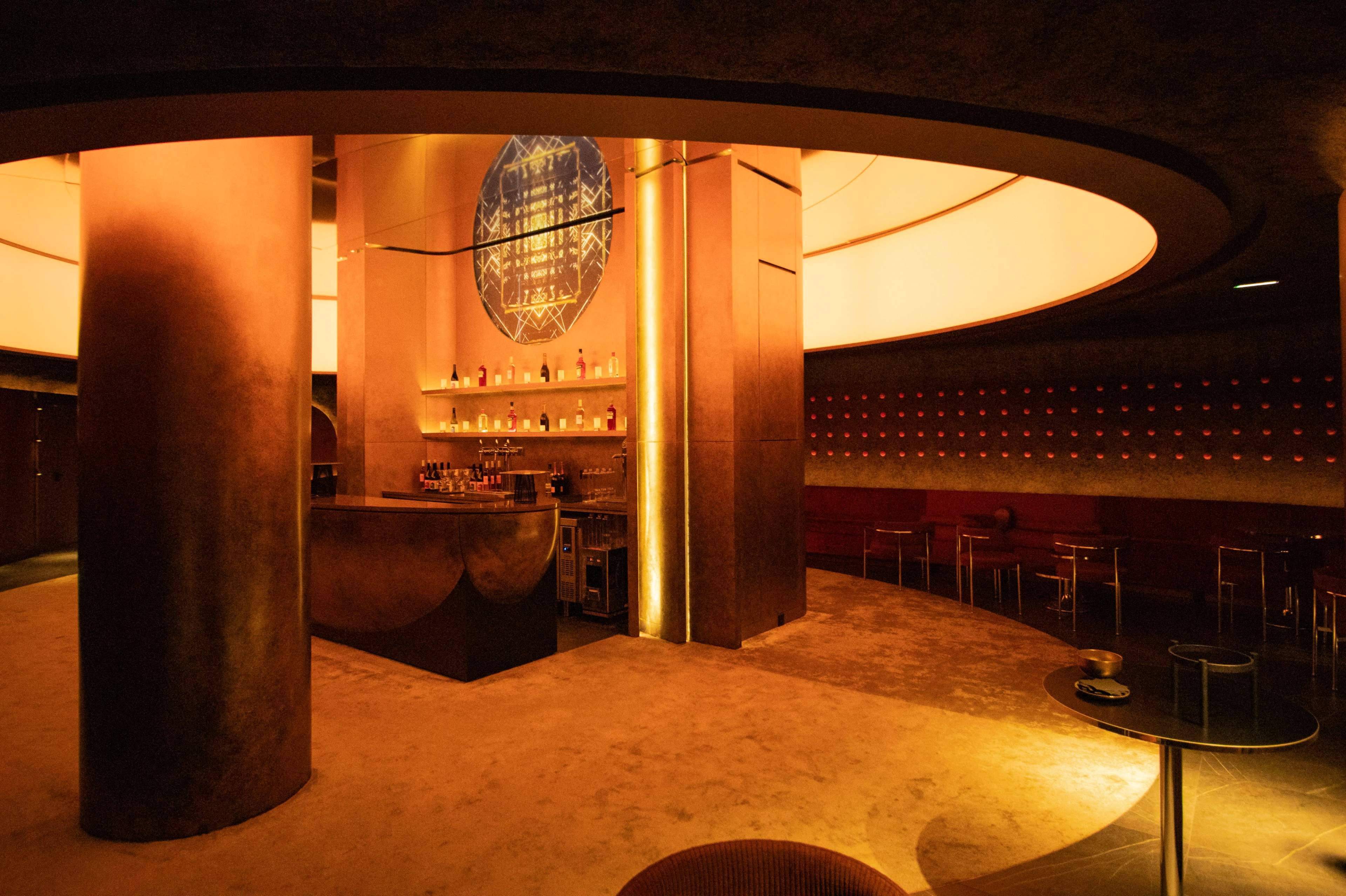 The image shows a modern bar interior with warm lighting, circular architectural elements, and a centrally located bar counter.