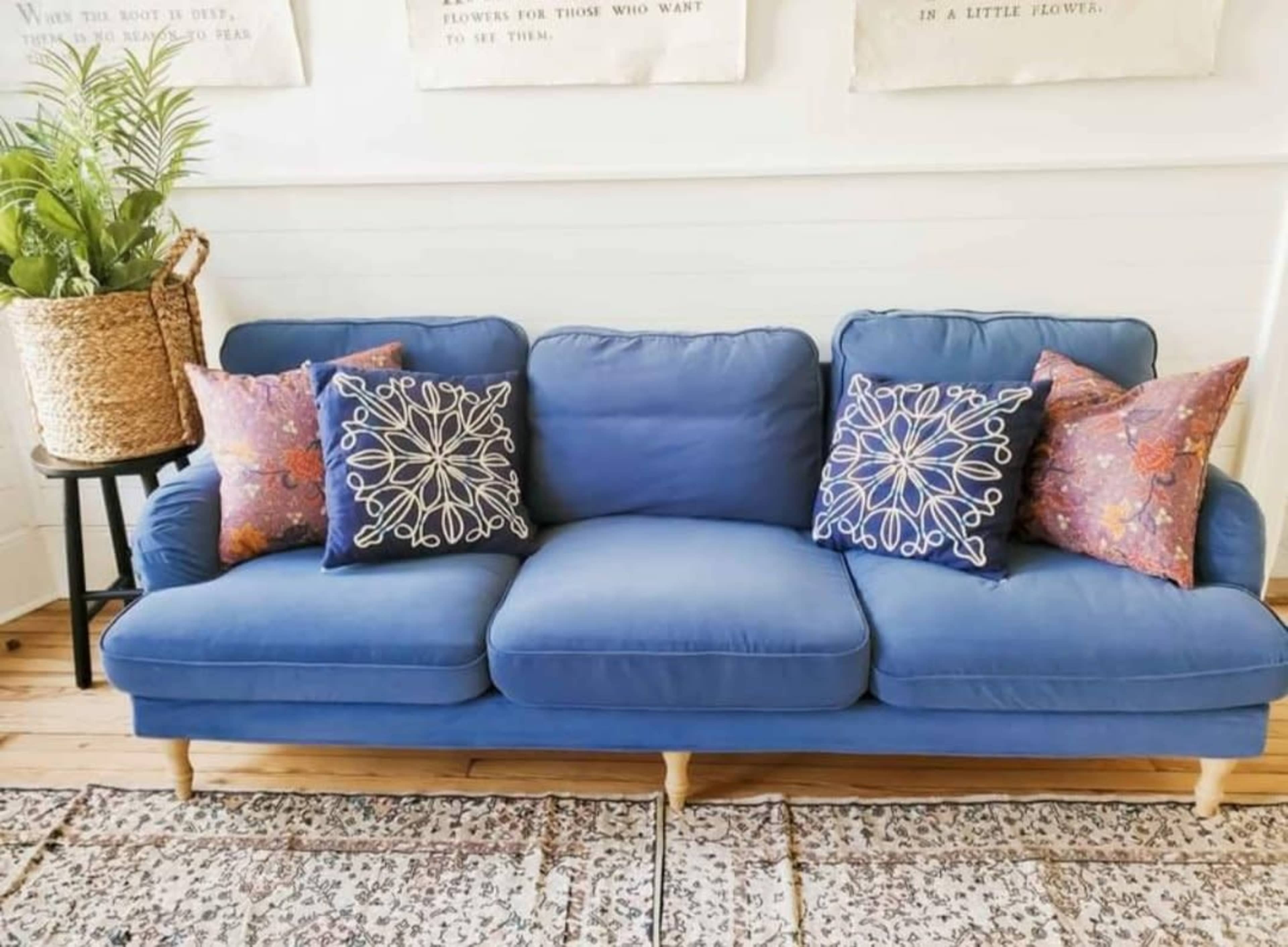 A blue sofa with five decorative pillows is positioned in a well-lit room with a plant and a patterned rug.